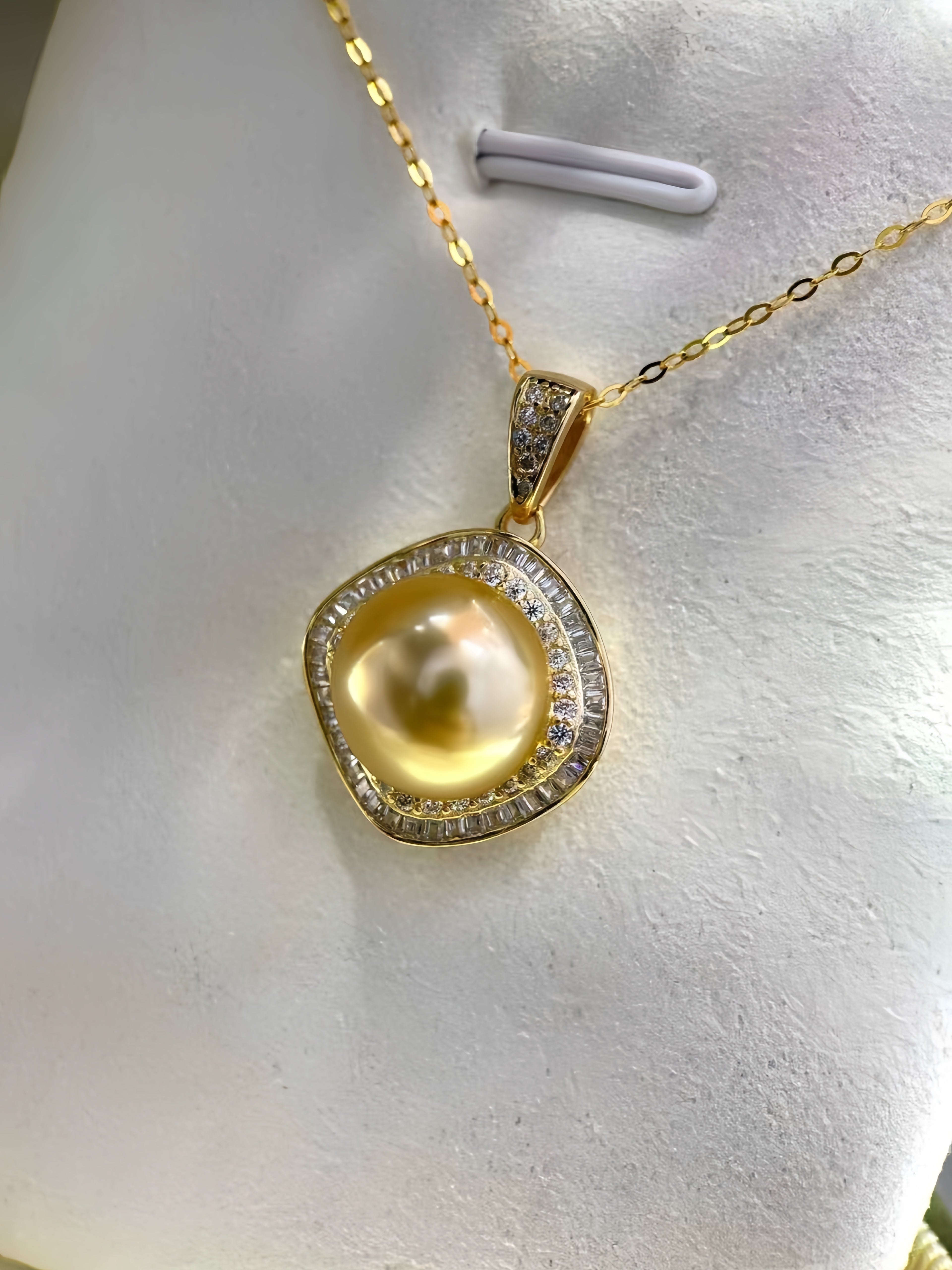 A gold pendant necklace featuring a large, lustrous yellow pearl surrounded by a halo of small clear stones, displayed on a white jewelry bust.