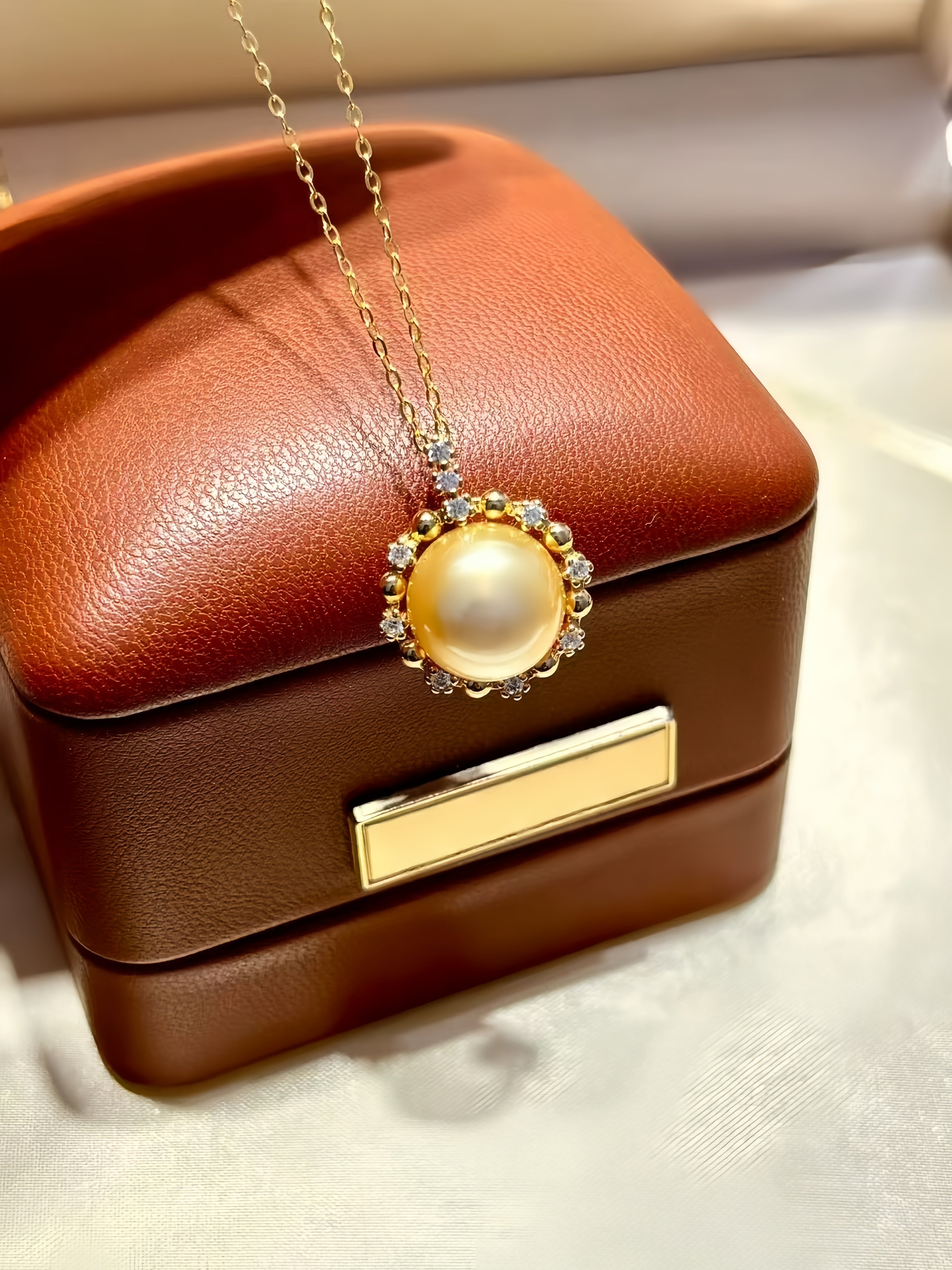 A gold pendant necklace with a large white pearl surrounded by small clear stones, displayed on a brown leather jewelry box.