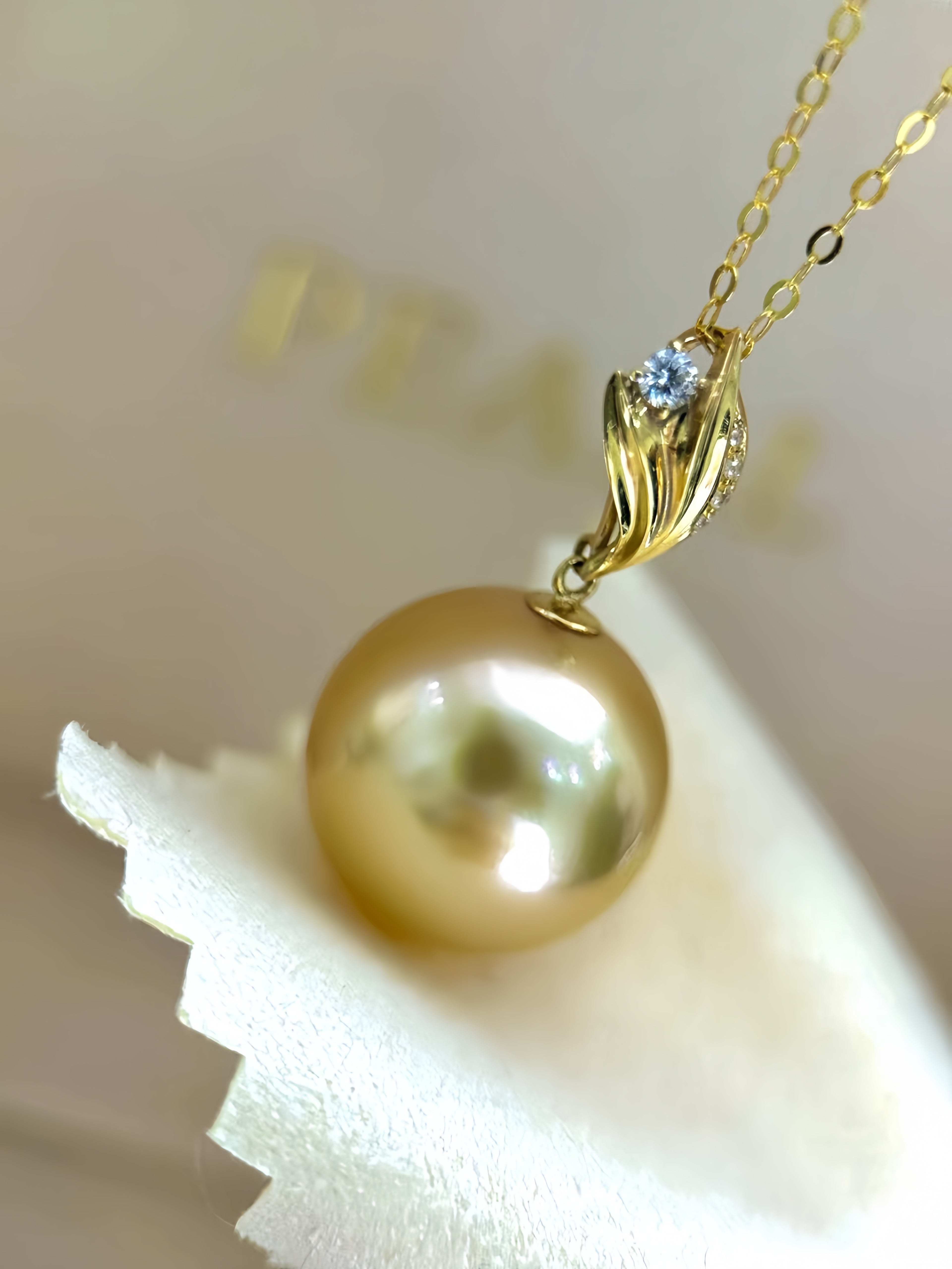 A gold pendant necklace featuring a large, lustrous gold pearl and a small clear gemstone, resting on a white textured surface.
