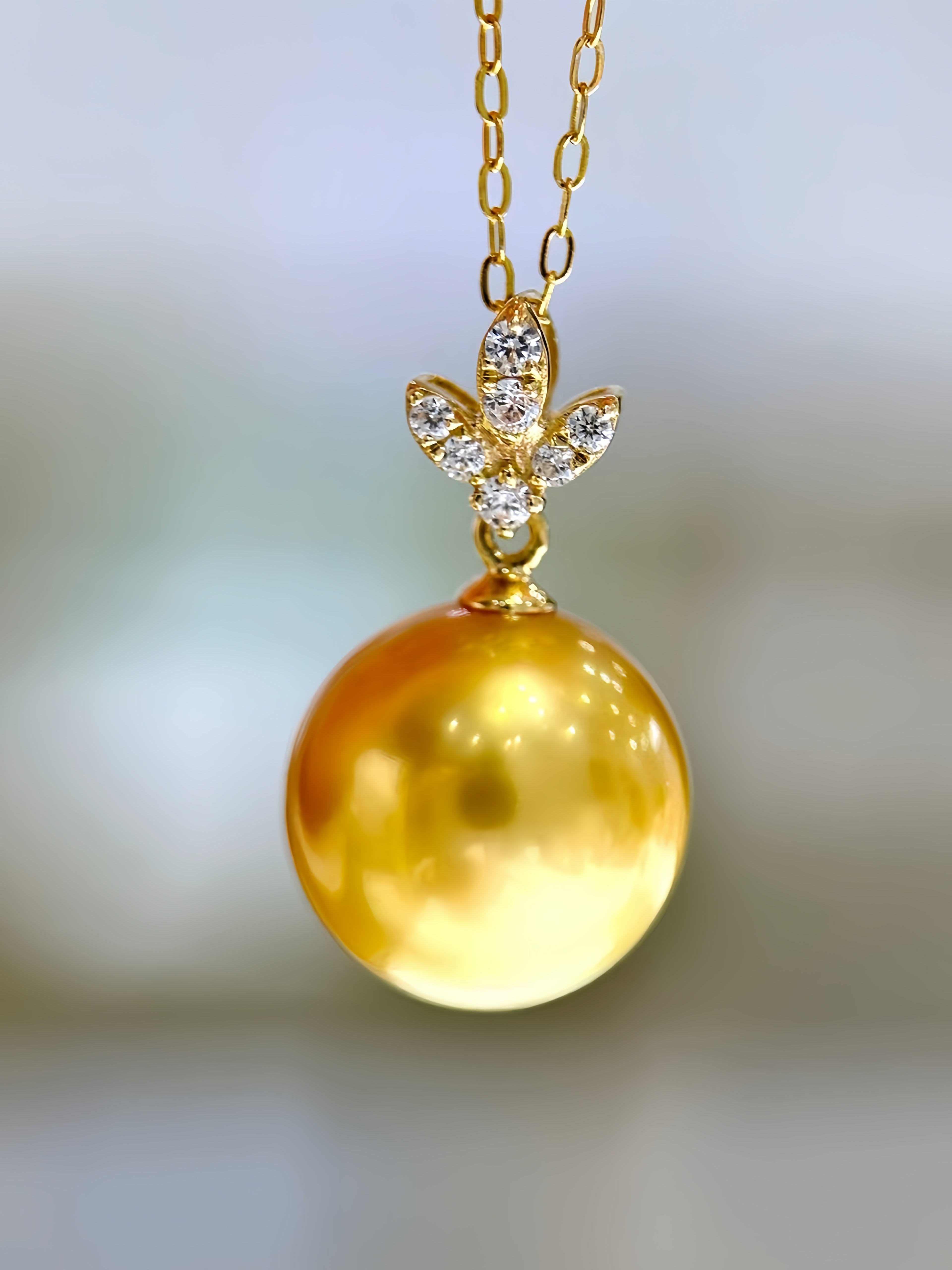 A gold pendant necklace featuring a large, round, lustrous golden-yellow pearl pendant and a delicate gold floral bail set with small clear stones, shown against a soft, out-of-focus light gray background.