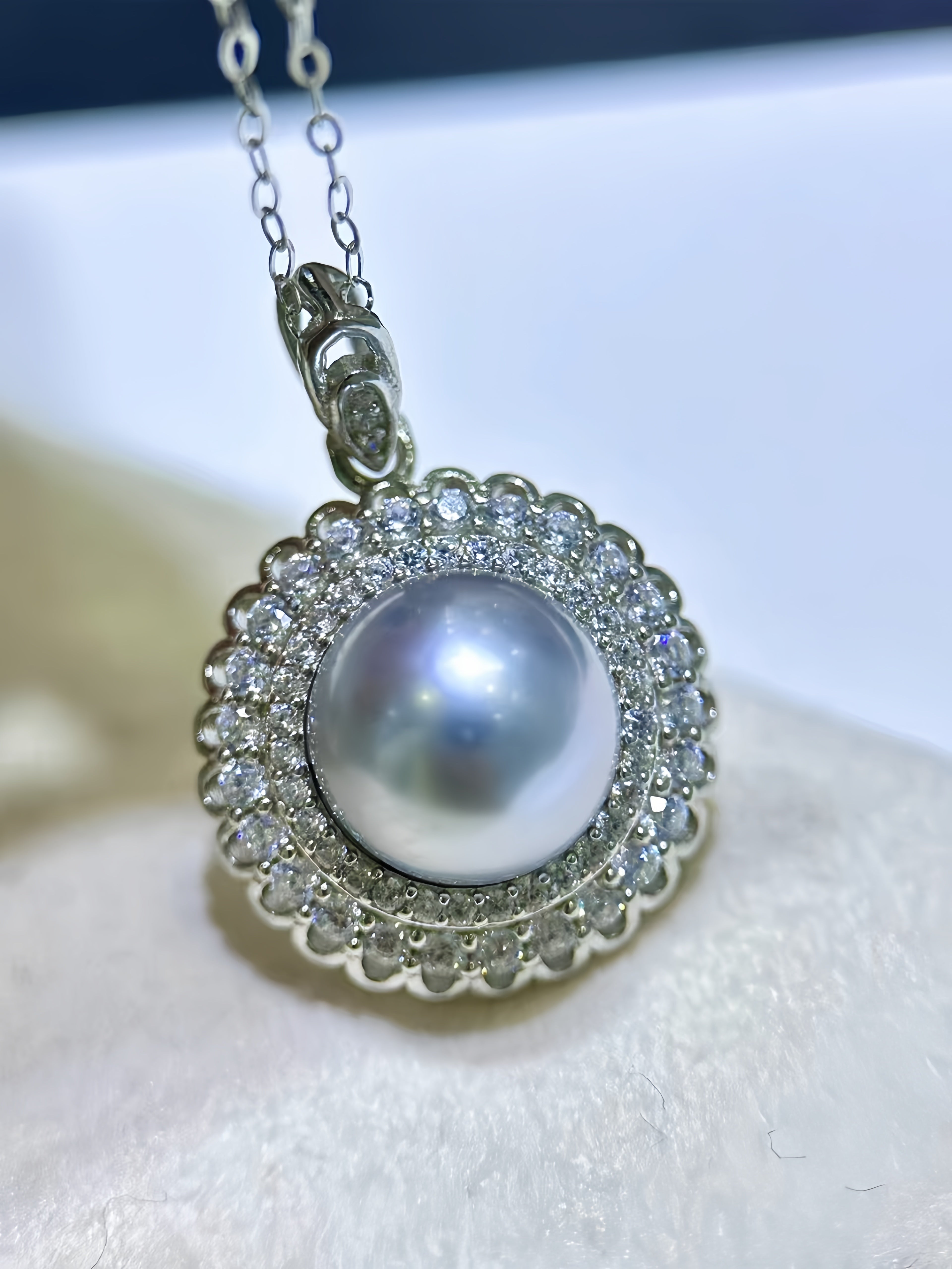 A round pearl pendant necklace with a halo of small clear stones, shown close up on a soft white surface. The pendant is attached to a silver-toned chain.