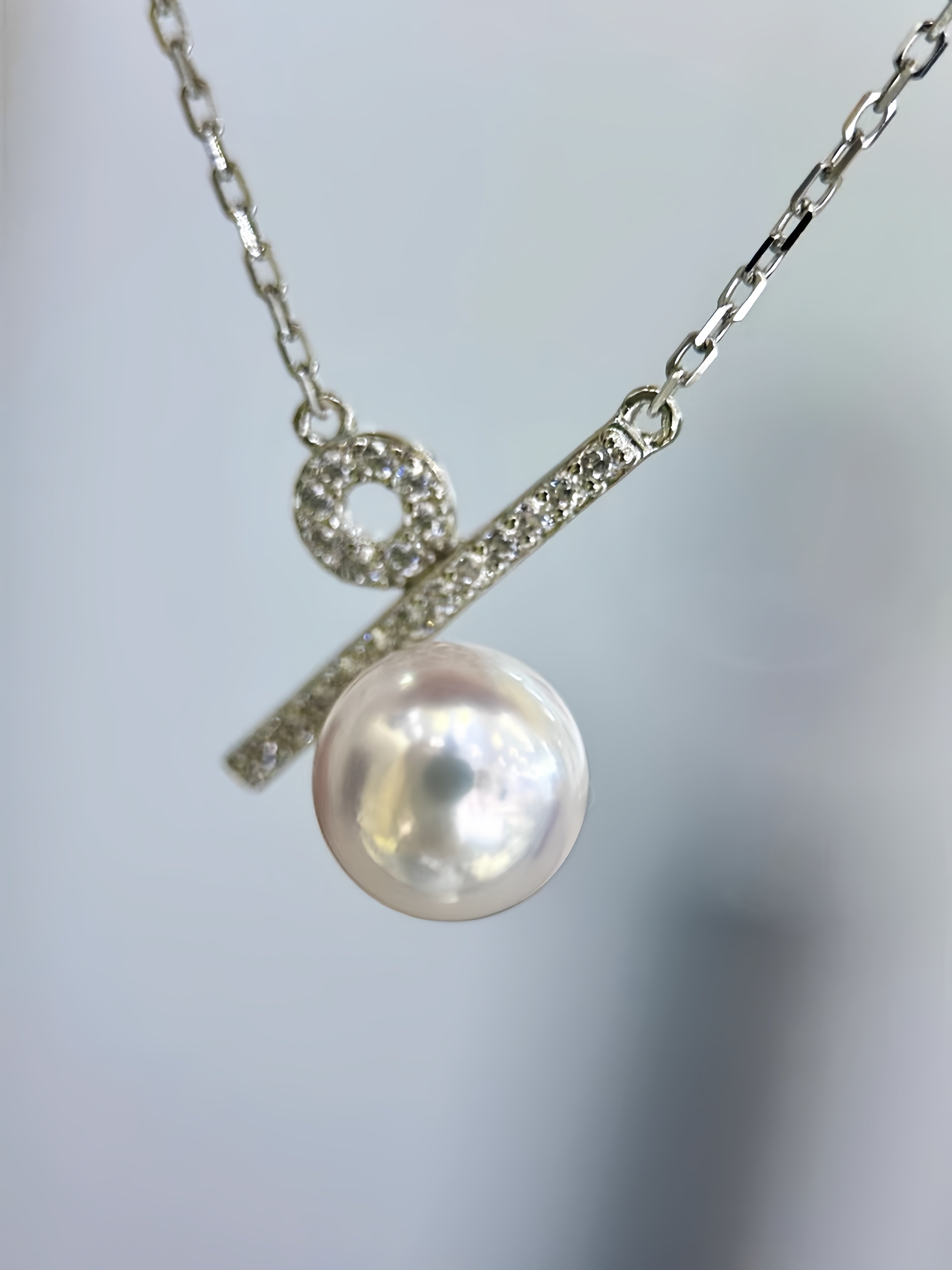 A silver-toned necklace featuring a bar pendant with a small round pavé detail and a dangling lustrous pearl, shown against a soft blurred light gray background.