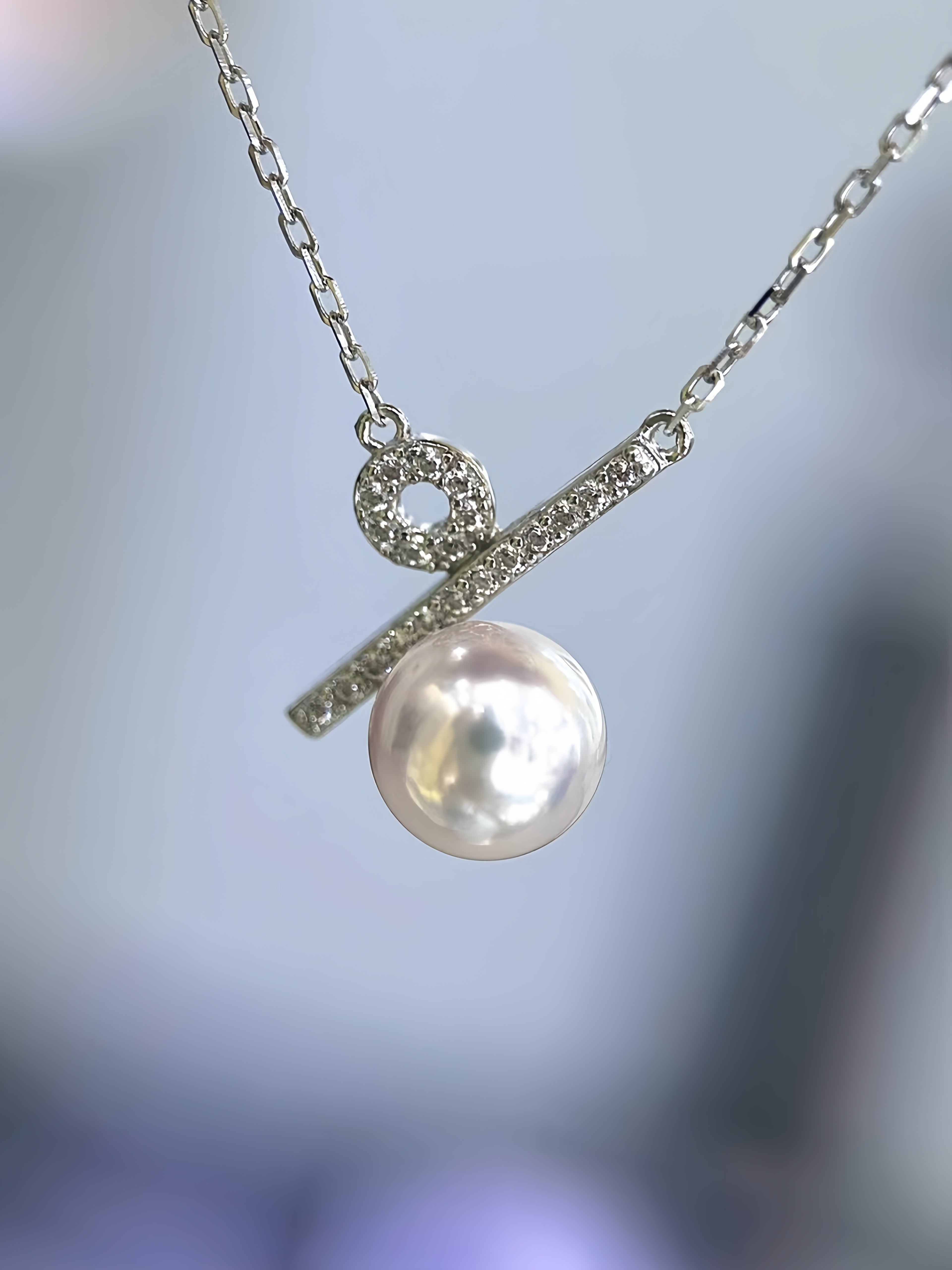 A silver necklace featuring a bar pendant with a single large, lustrous white pearl and a small round pavé detail, shown against a soft, blurred blue-gray background.
