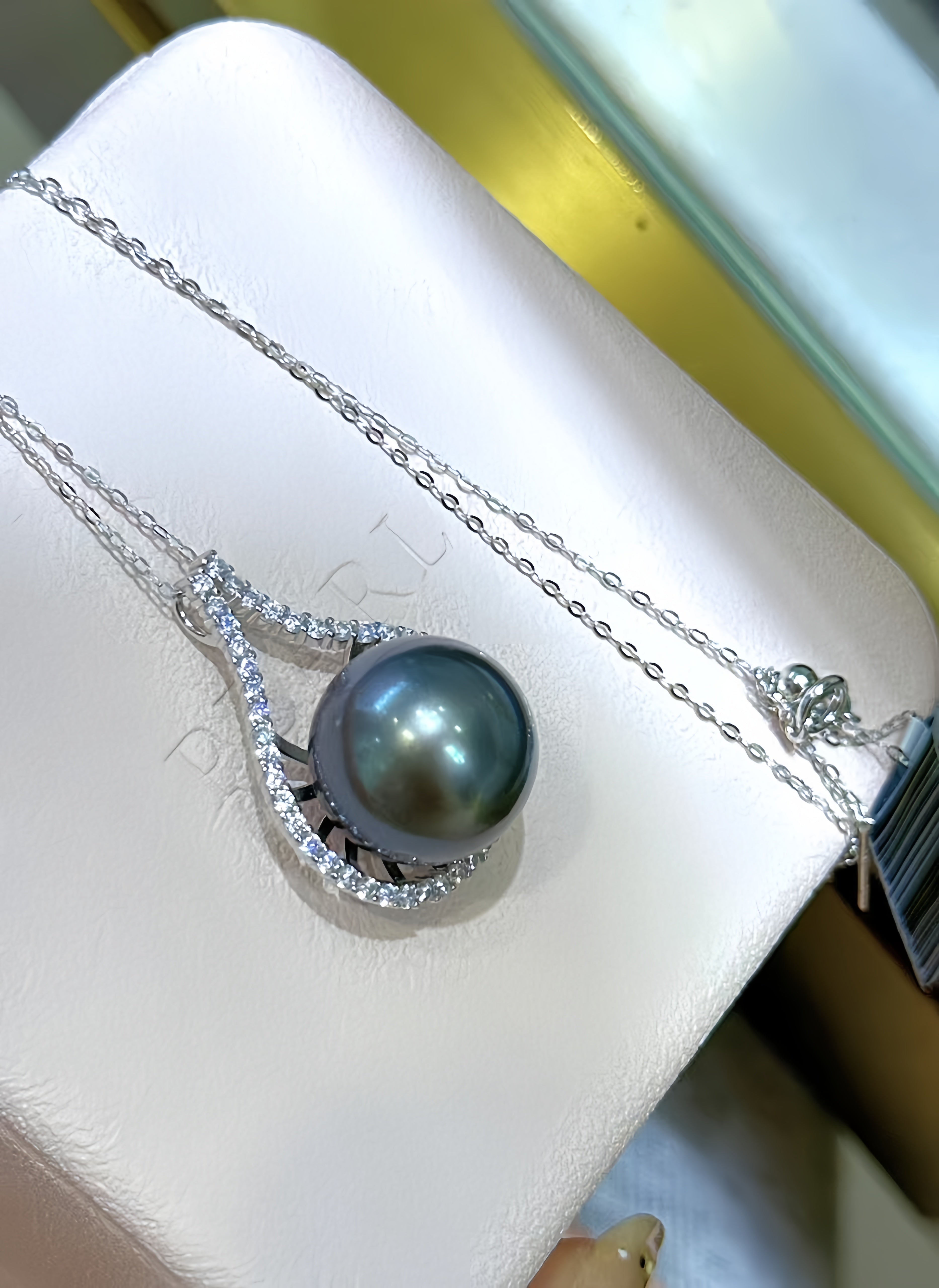 A silver necklace with a teardrop-shaped pendant featuring a dark gray pearl and small clear stones, displayed on a white jewelry box.