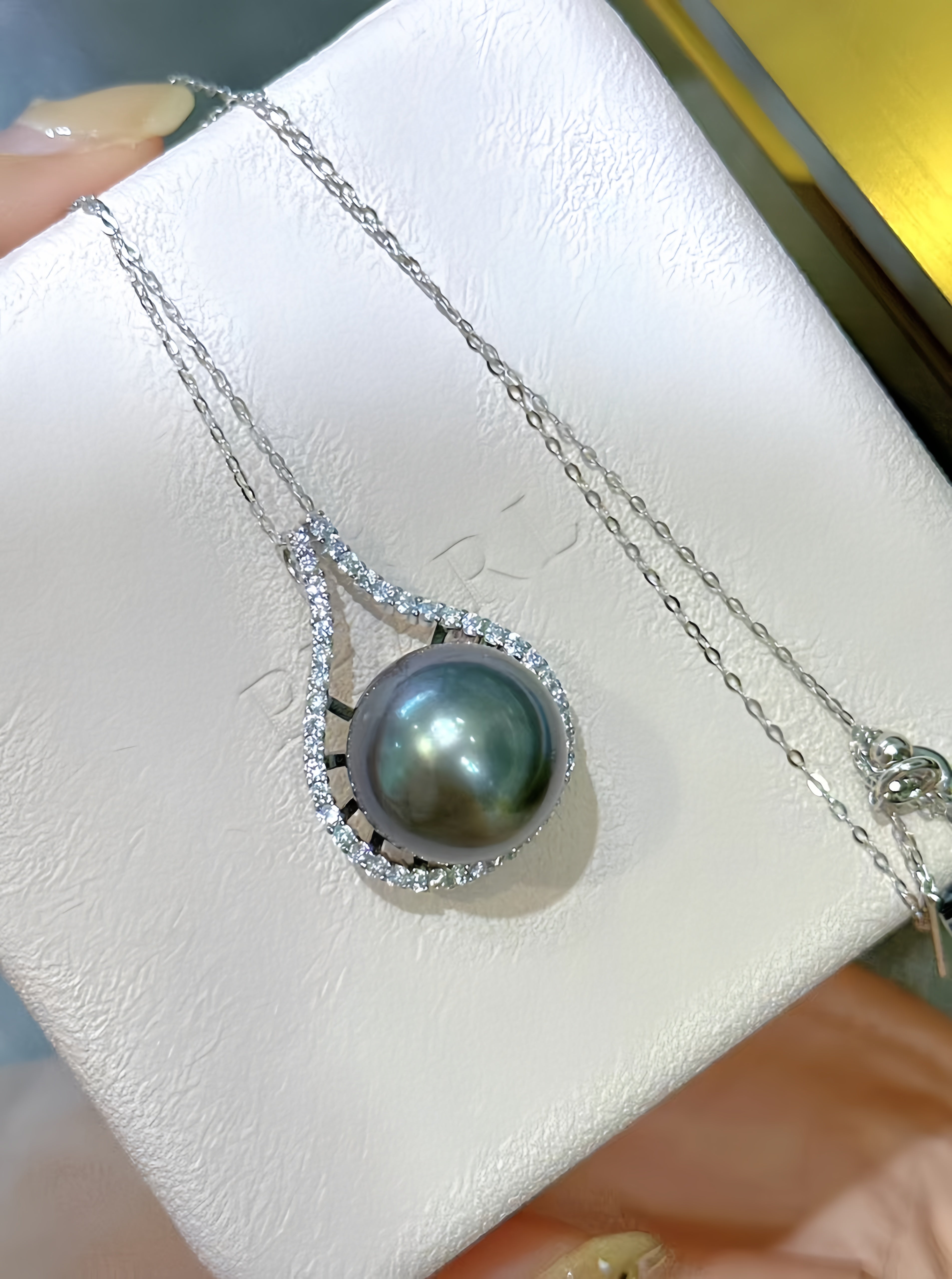 A silver teardrop pendant necklace featuring a dark green-gray Tahitian pearl surrounded by a halo of small clear stones, displayed on a textured white jewelry box.