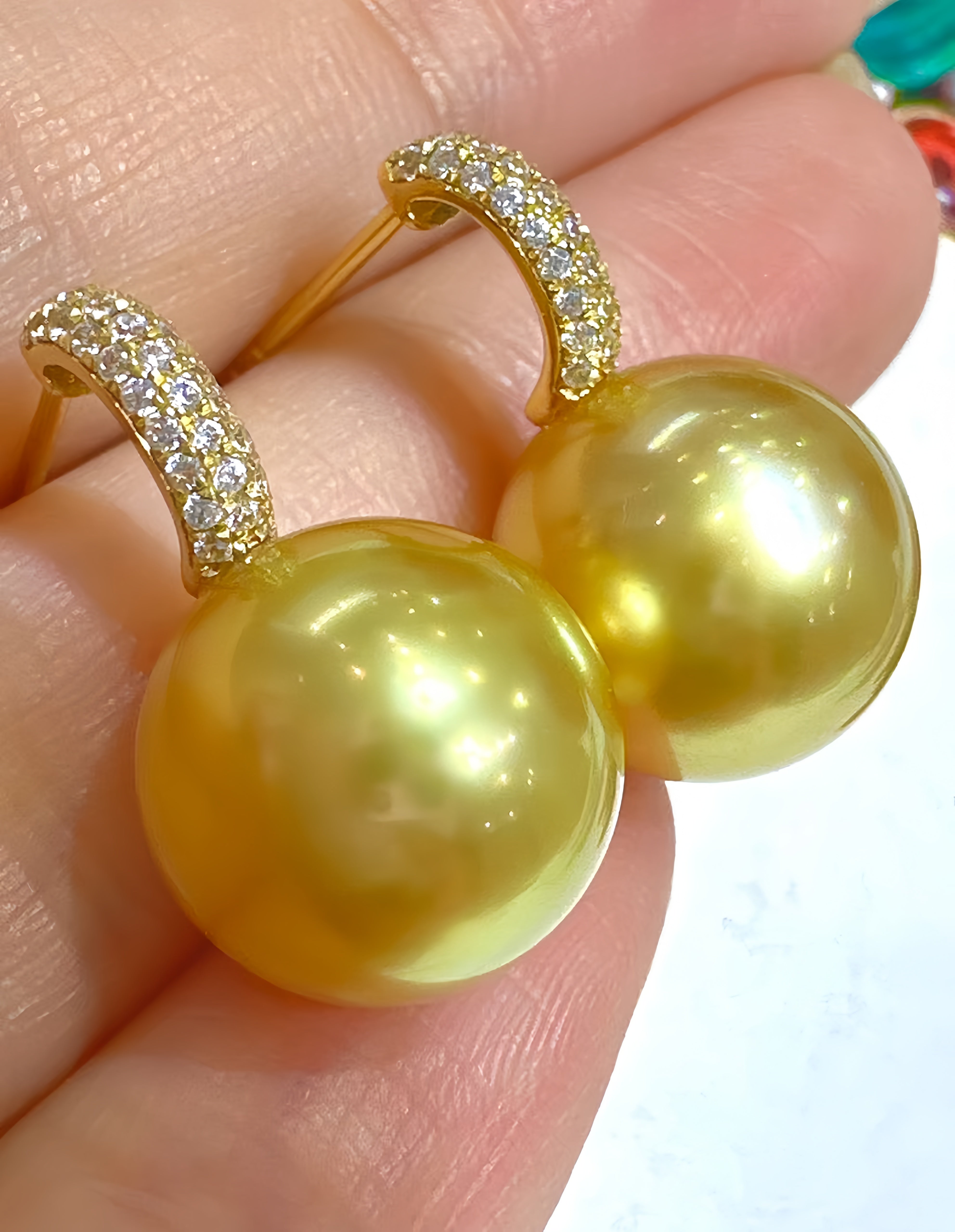 A pair of gold hoop earrings with large round yellow-gold pearl-like drops and rows of small clear stones along the hoops, shown resting on a person's fingers.