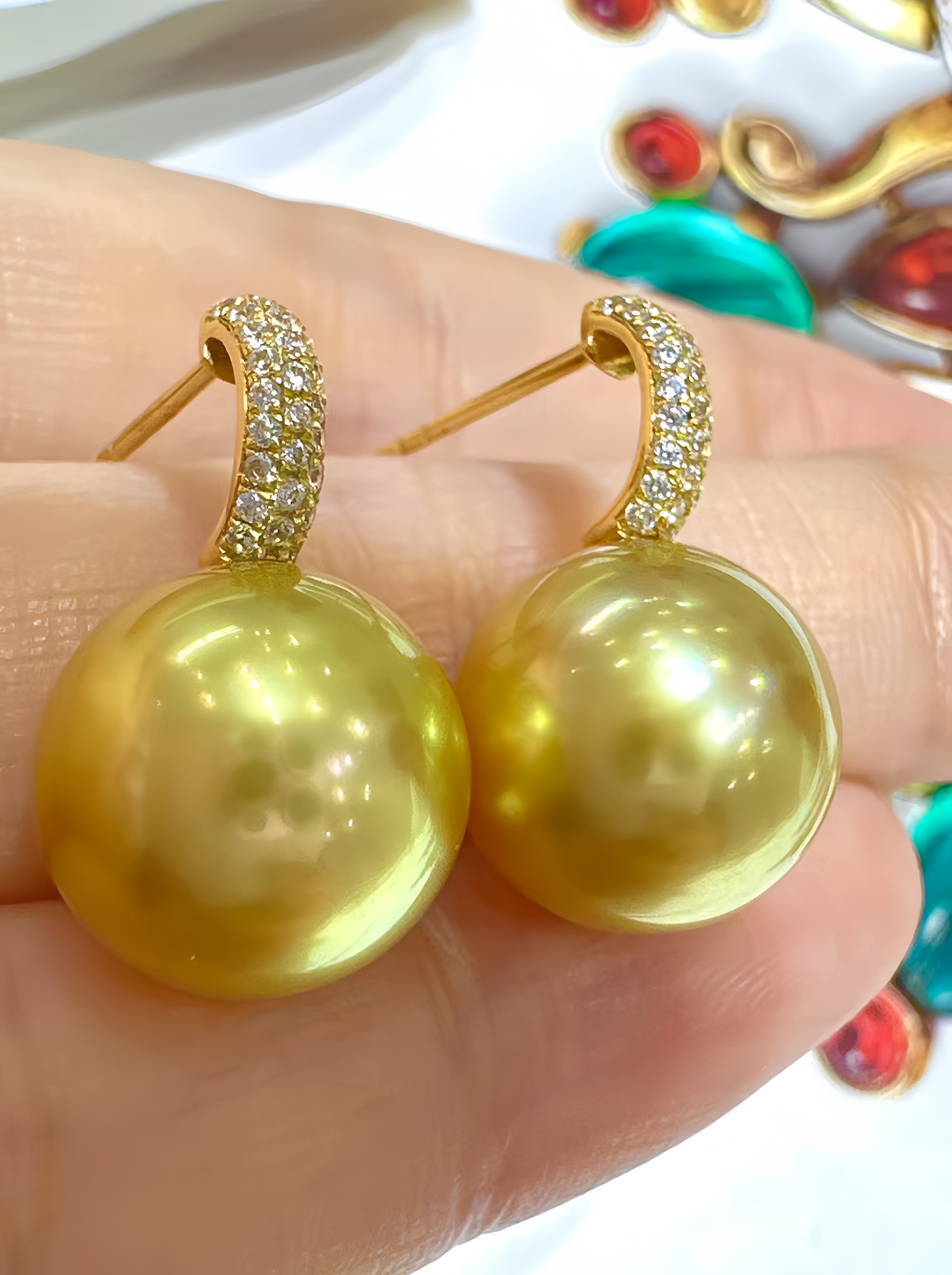 A pair of gold hoop earrings with large round yellow-gold pearl drops, held on a person's fingers. The hoops are set with small clear stones.