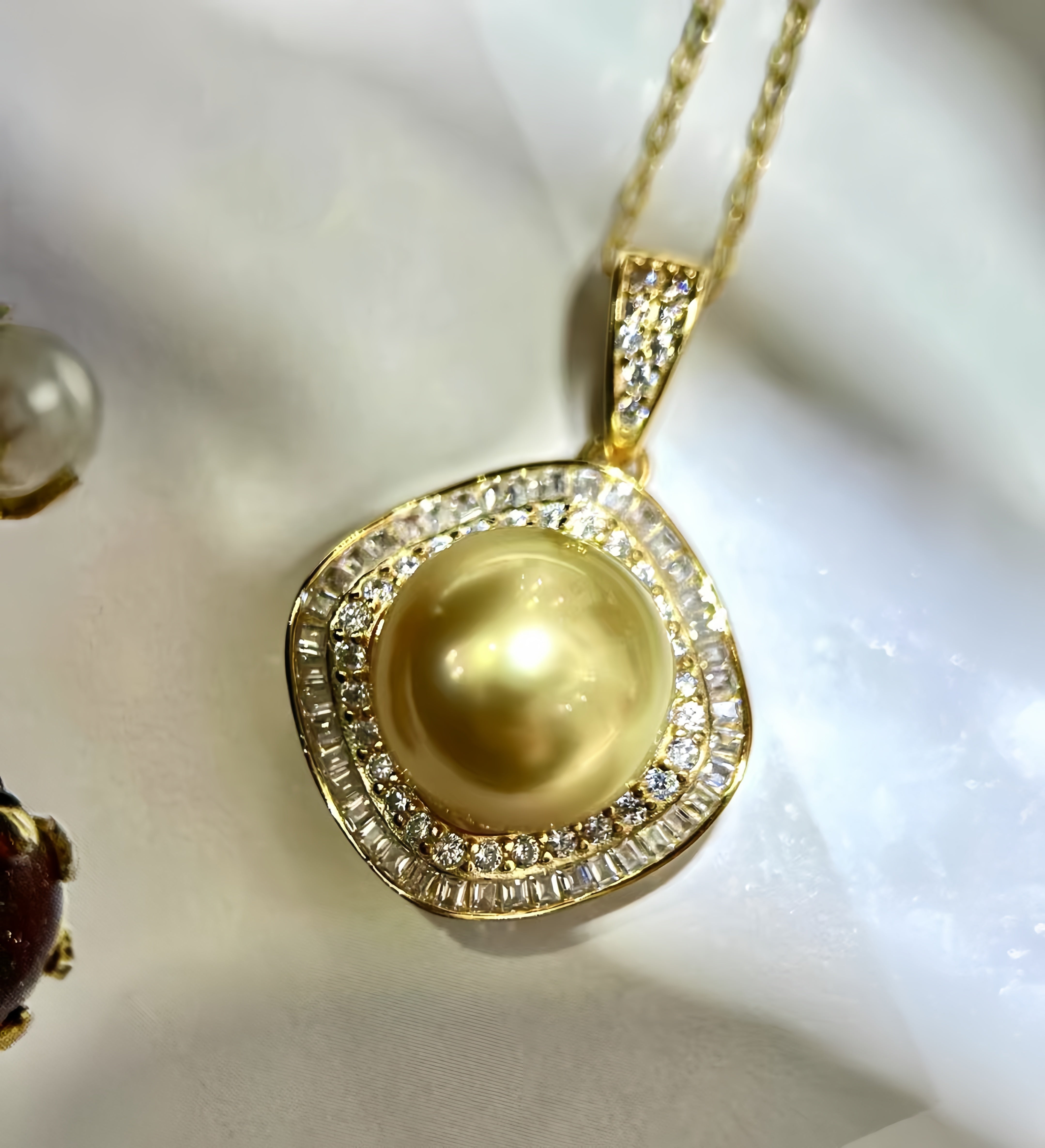 A gold pendant necklace featuring a large round yellow pearl surrounded by a halo of small clear stones, displayed on a soft white fabric background.