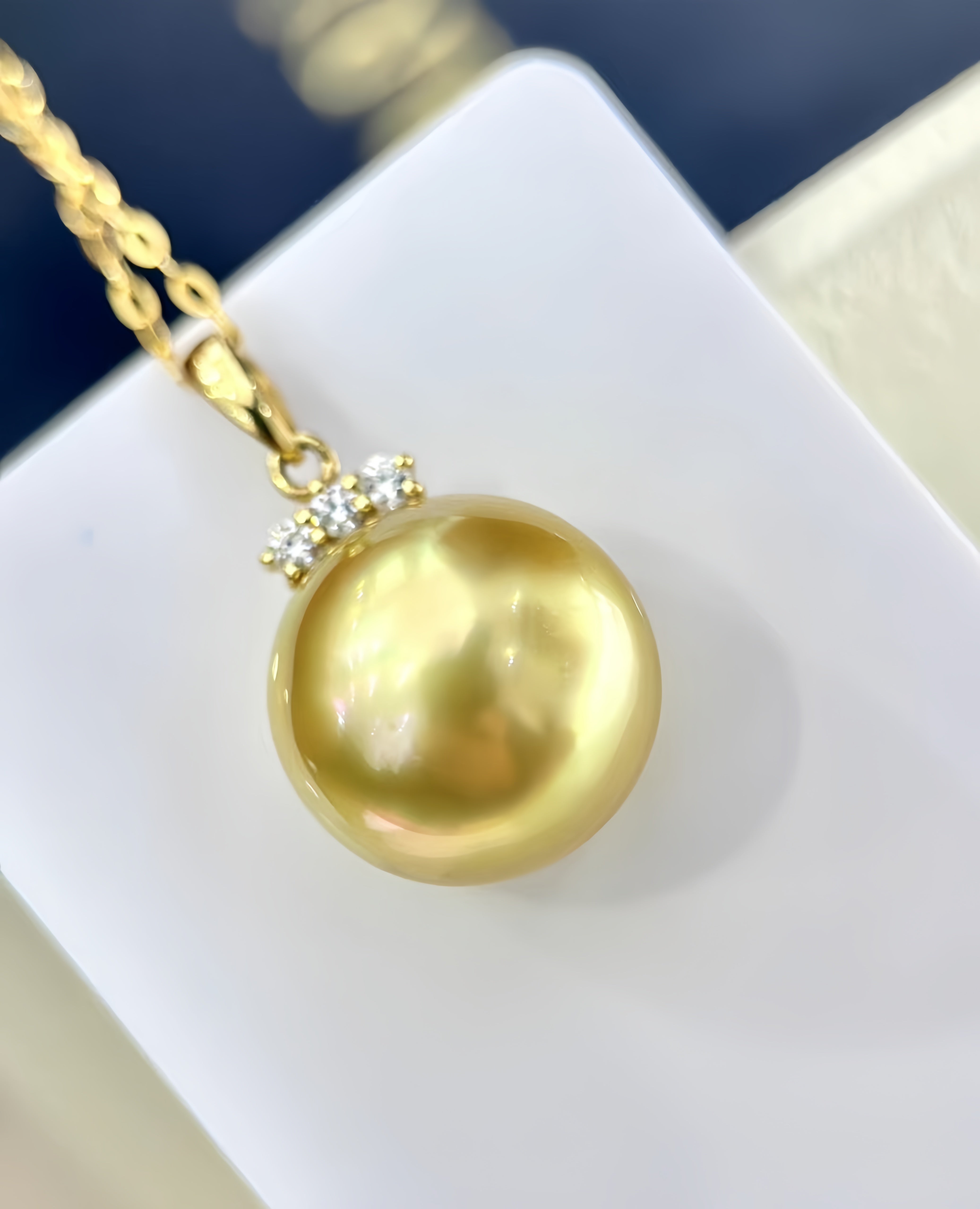 A gold pendant necklace featuring a large, lustrous gold freshwater pearl accented by small clear stones at the bail, resting on a white display block.