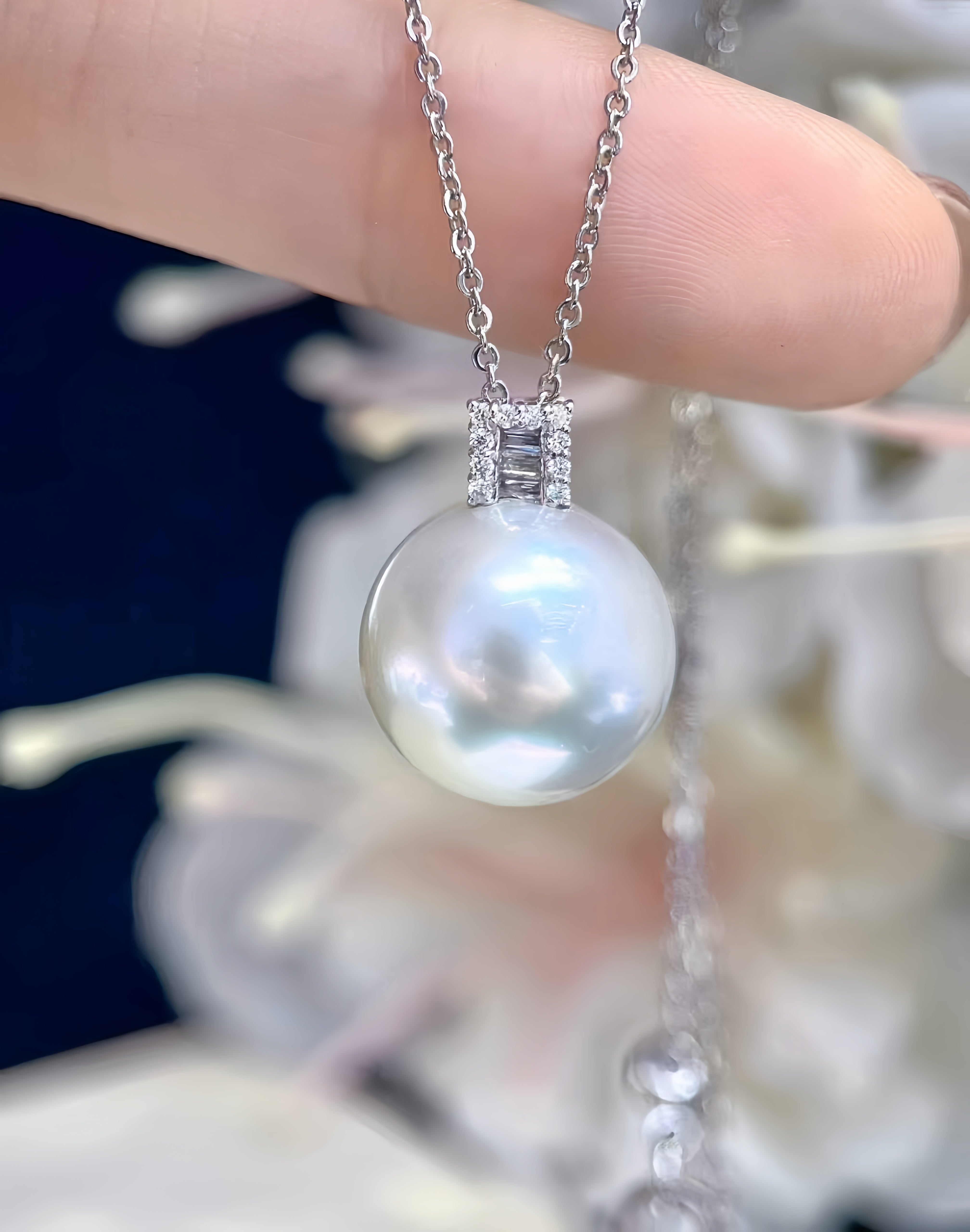 A close-up of a silver chain necklace with a large, lustrous white pearl pendant held by a person's fingers, with a blurred background of white flowers and a dark blue surface.