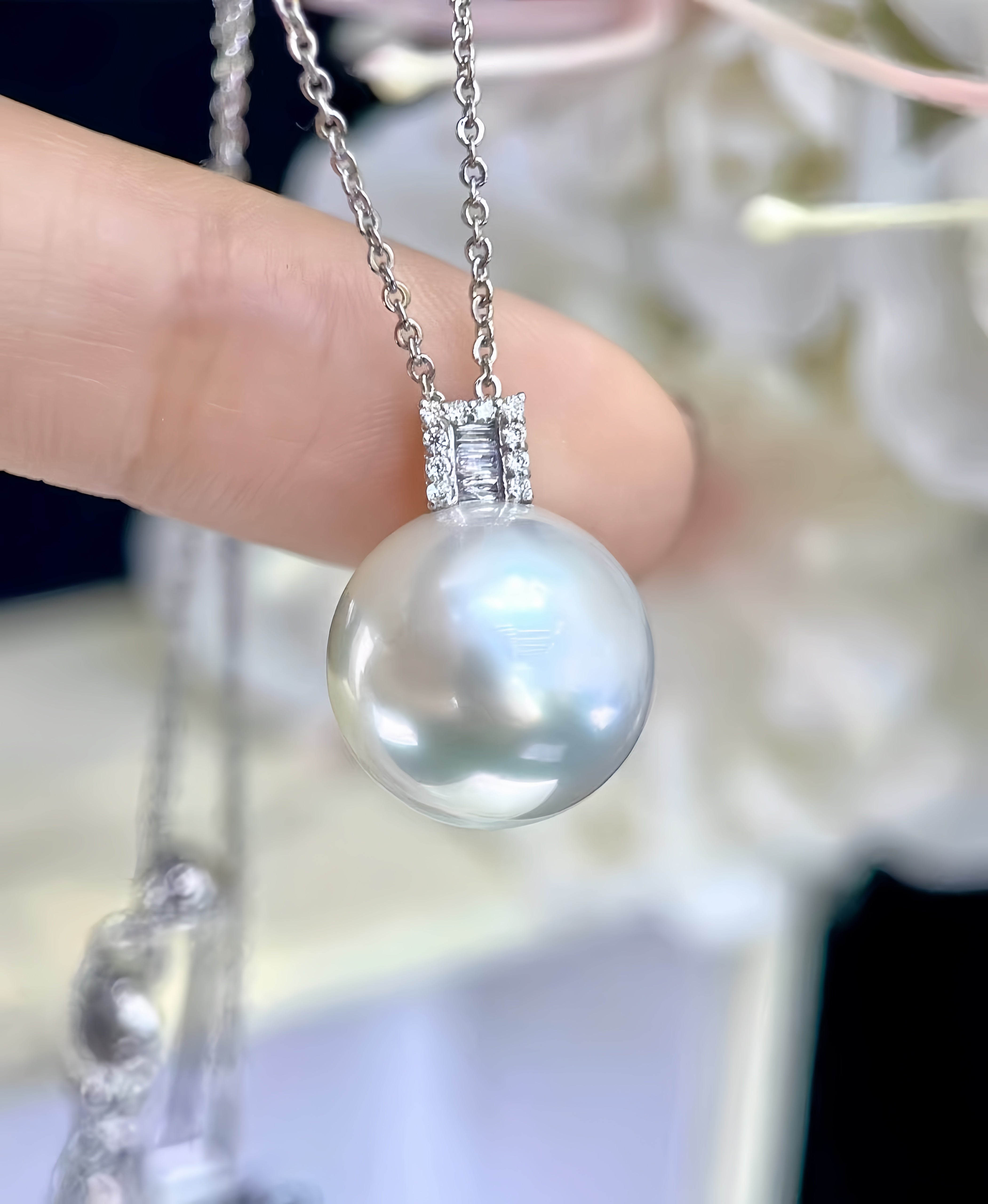 A close-up of a silver chain necklace with a large, round, lustrous white pearl pendant held by a person's finger. The pendant is set in a silver bezel with small clear stones.