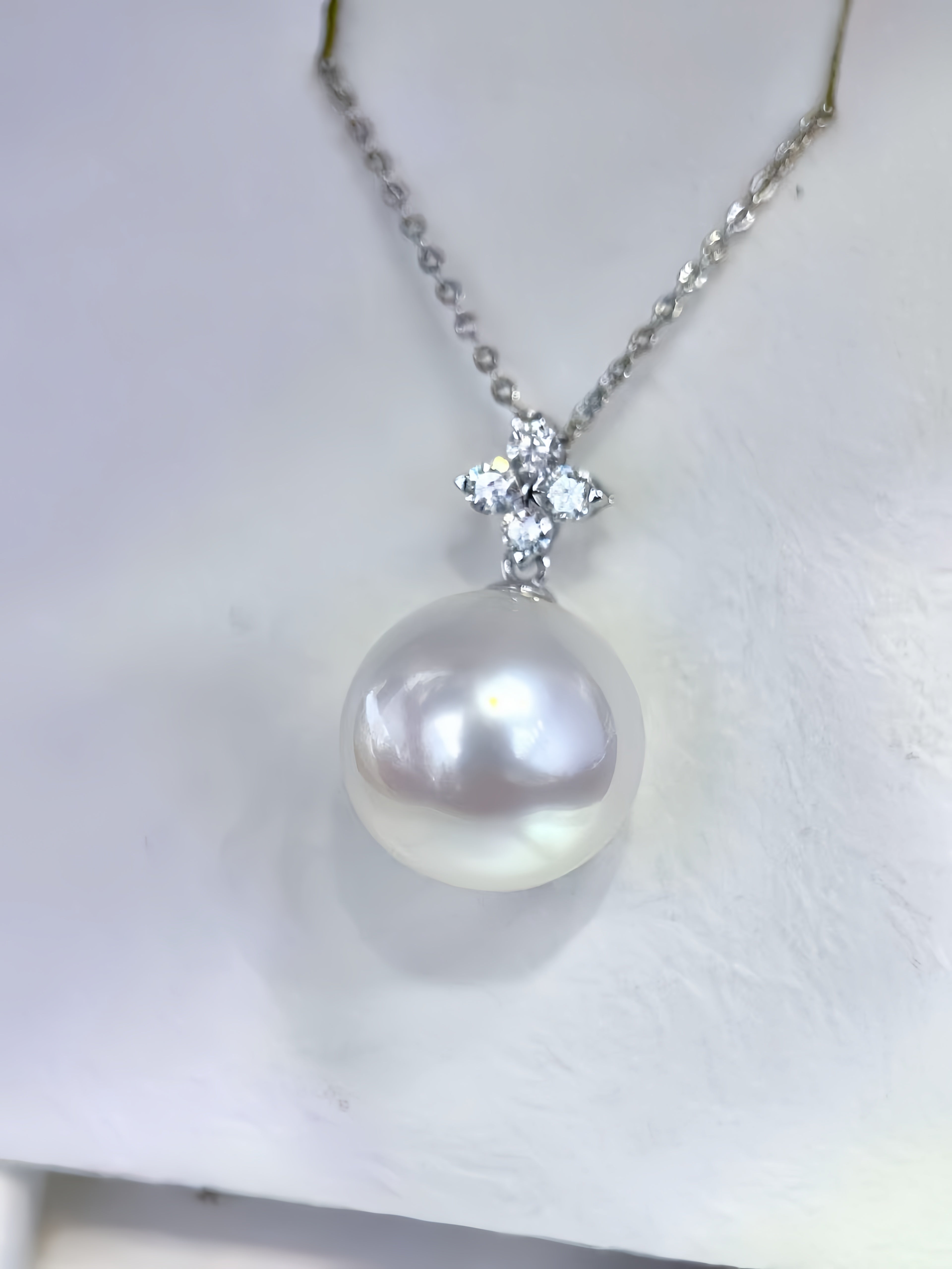 A silver chain necklace featuring a large, lustrous white pearl pendant with a sparkling clear crystal cross detail at the top, displayed on a textured white surface.