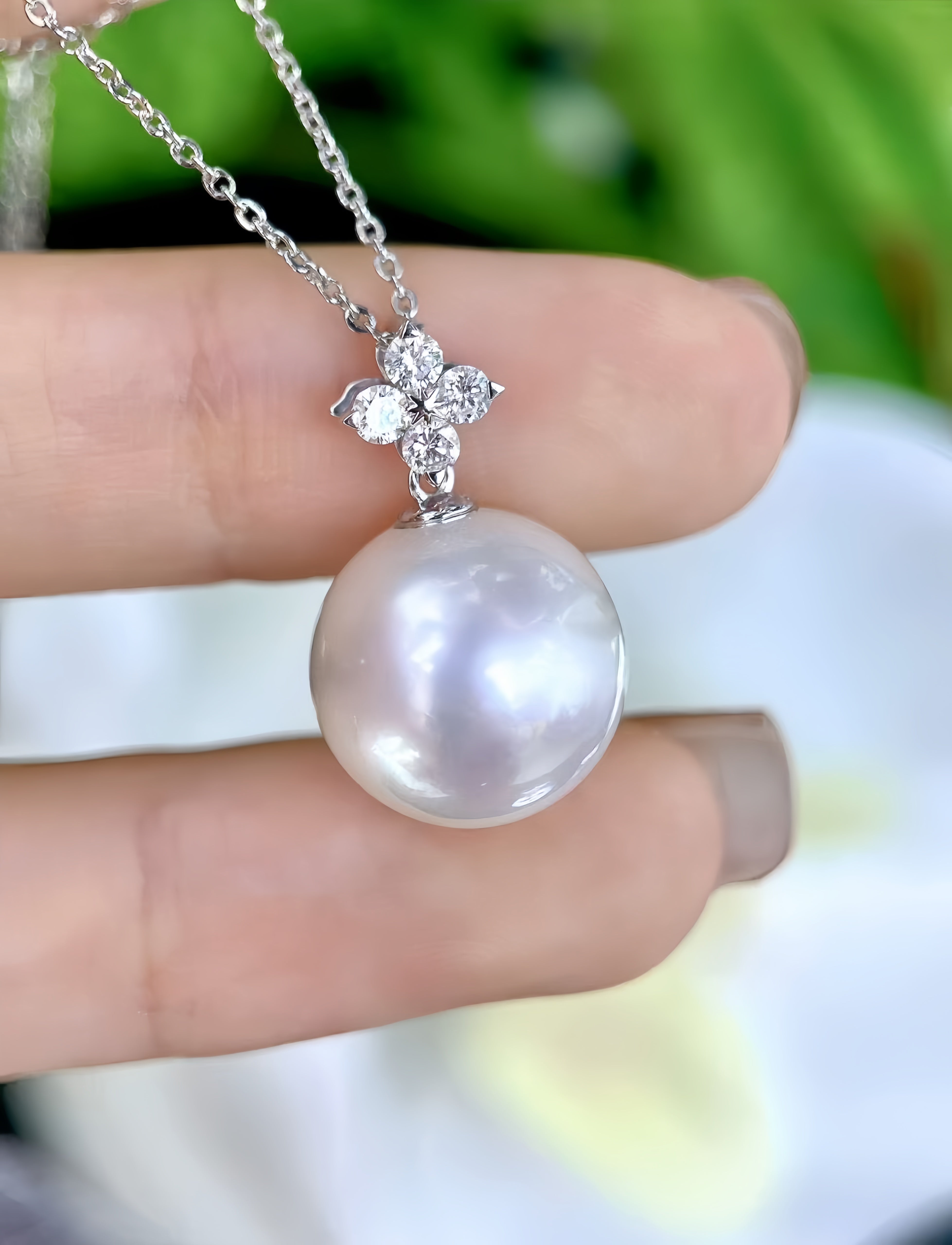 A close-up of a silver necklace with a large round white pearl pendant and a small floral cluster of clear stones, held between fingers against a blurred green and white background.