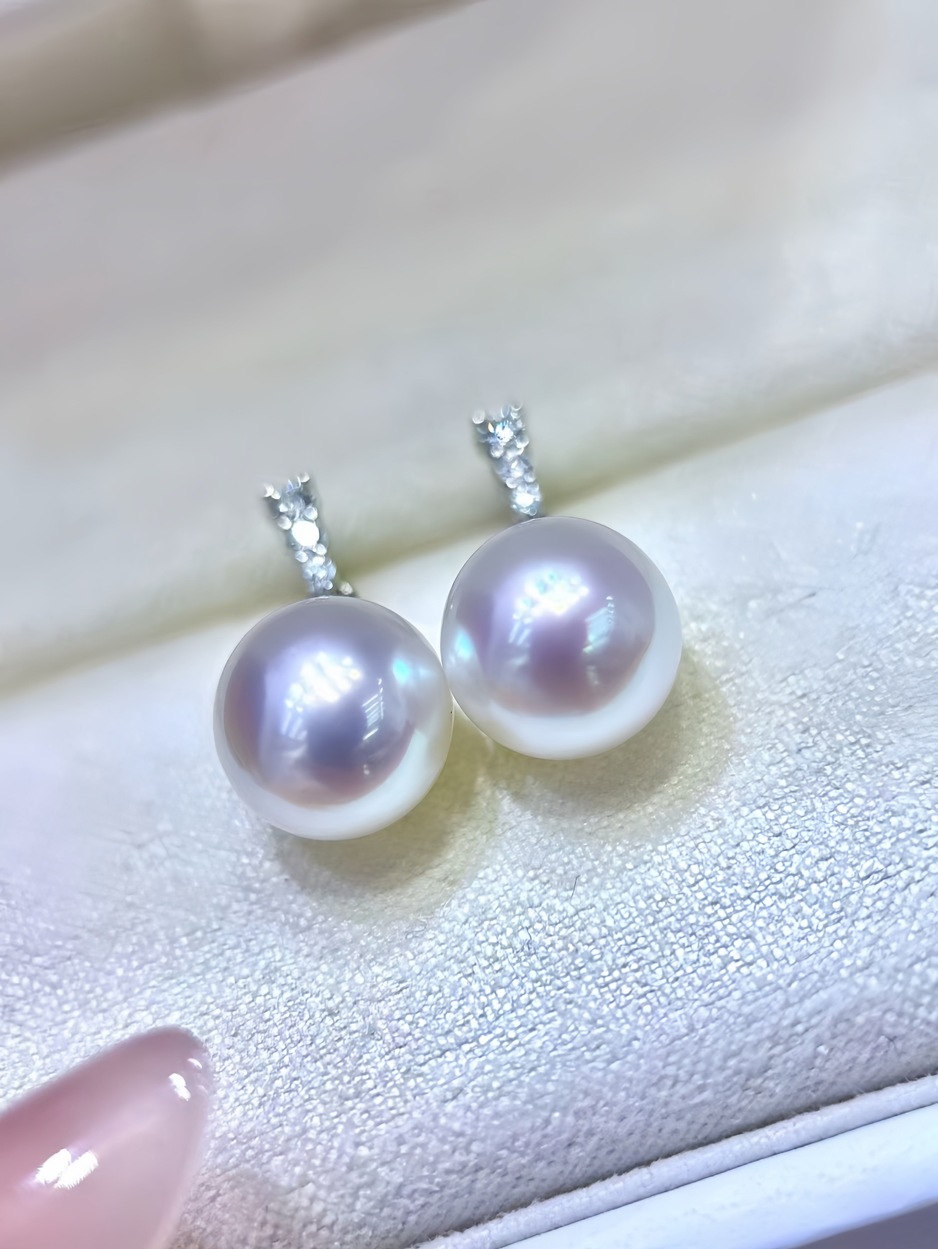 A pair of freshwater pearl stud earrings with silver-toned bezel-set clear stones, displayed on a white textured jewelry box.