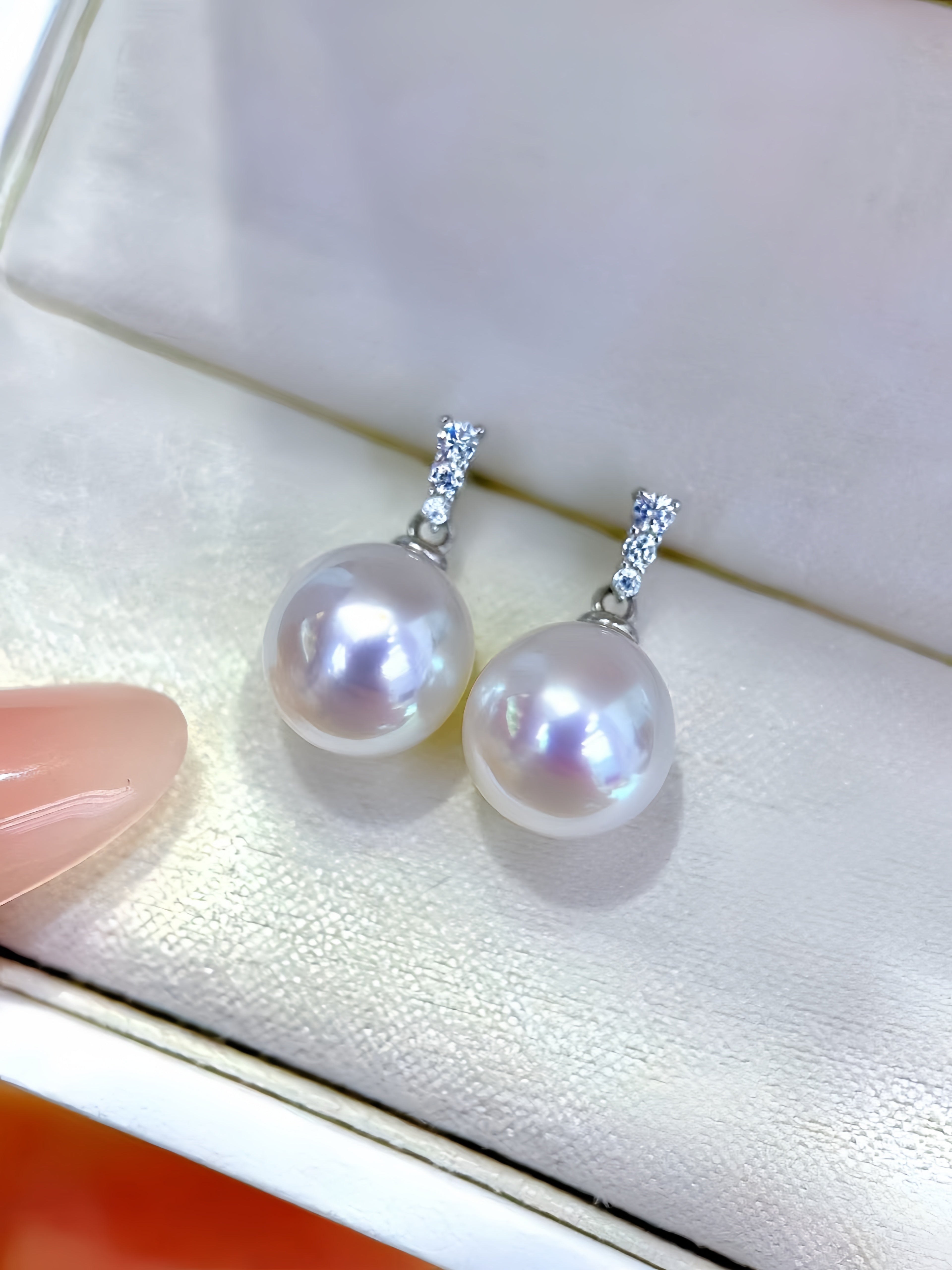 A pair of silver drop earrings with large lustrous white pearls and small clear stones at the top, displayed on a cream jewelry box. A finger with pink nail polish is partially visible at the left edge.