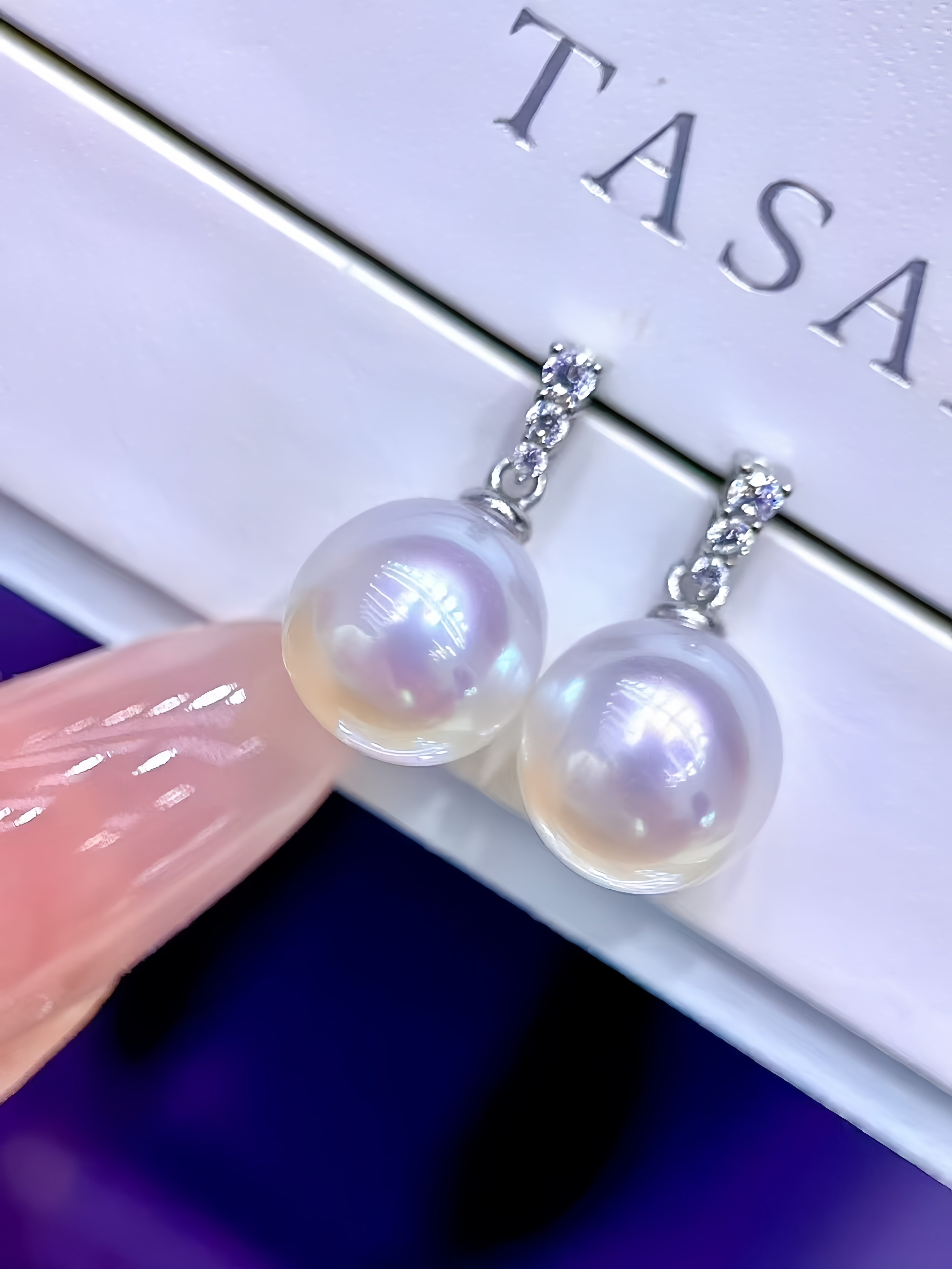 A close-up of a pair of dangling pearl earrings with silver-toned settings and small clear stones, held near a white box branded TASAR. A finger with pink manicured nails is visible at the lower left.