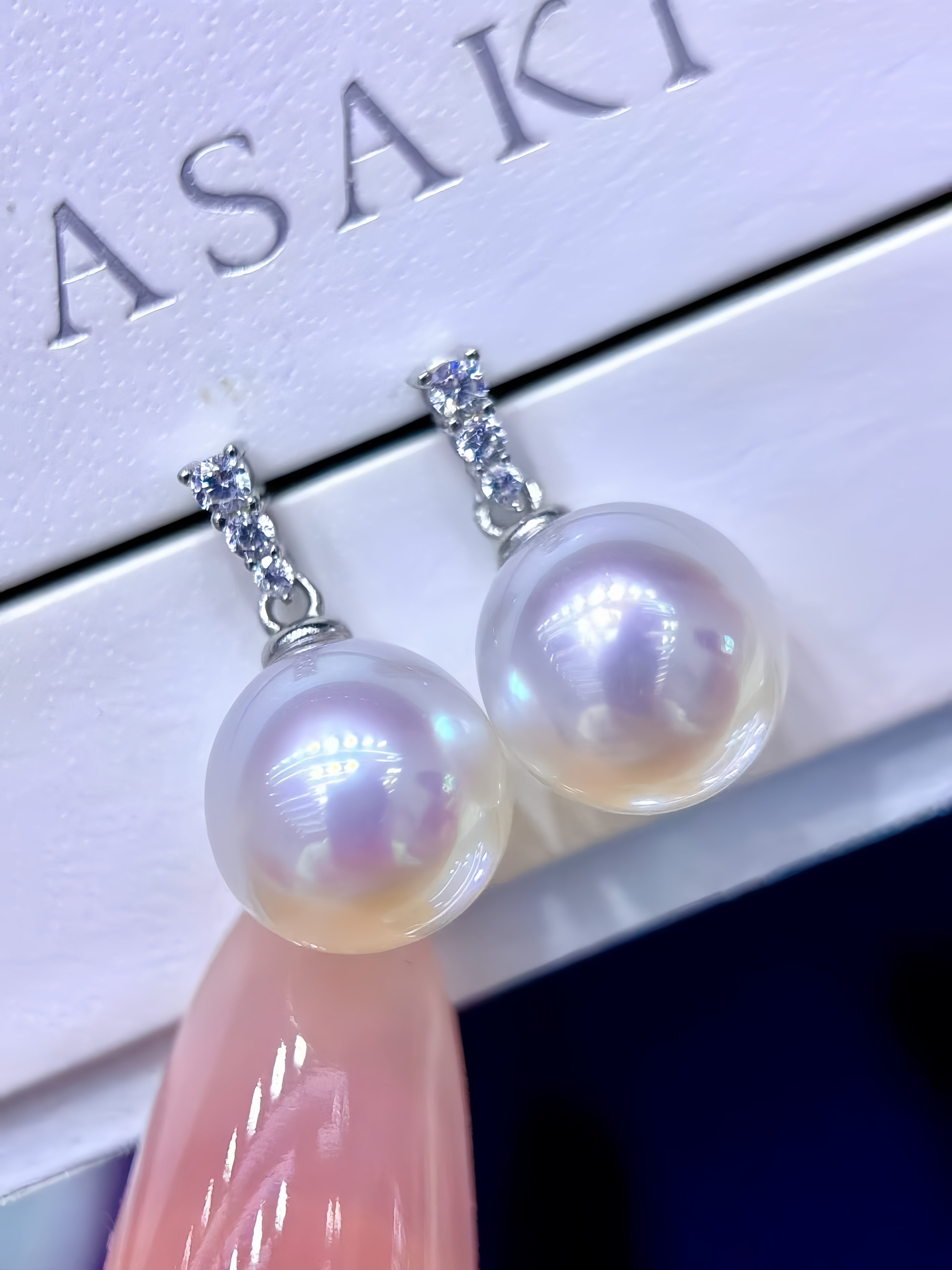 A pair of Asaki brand pearl drop earrings with silver-toned settings, resting on a white branded display box with a pink manicured finger pointing at them.