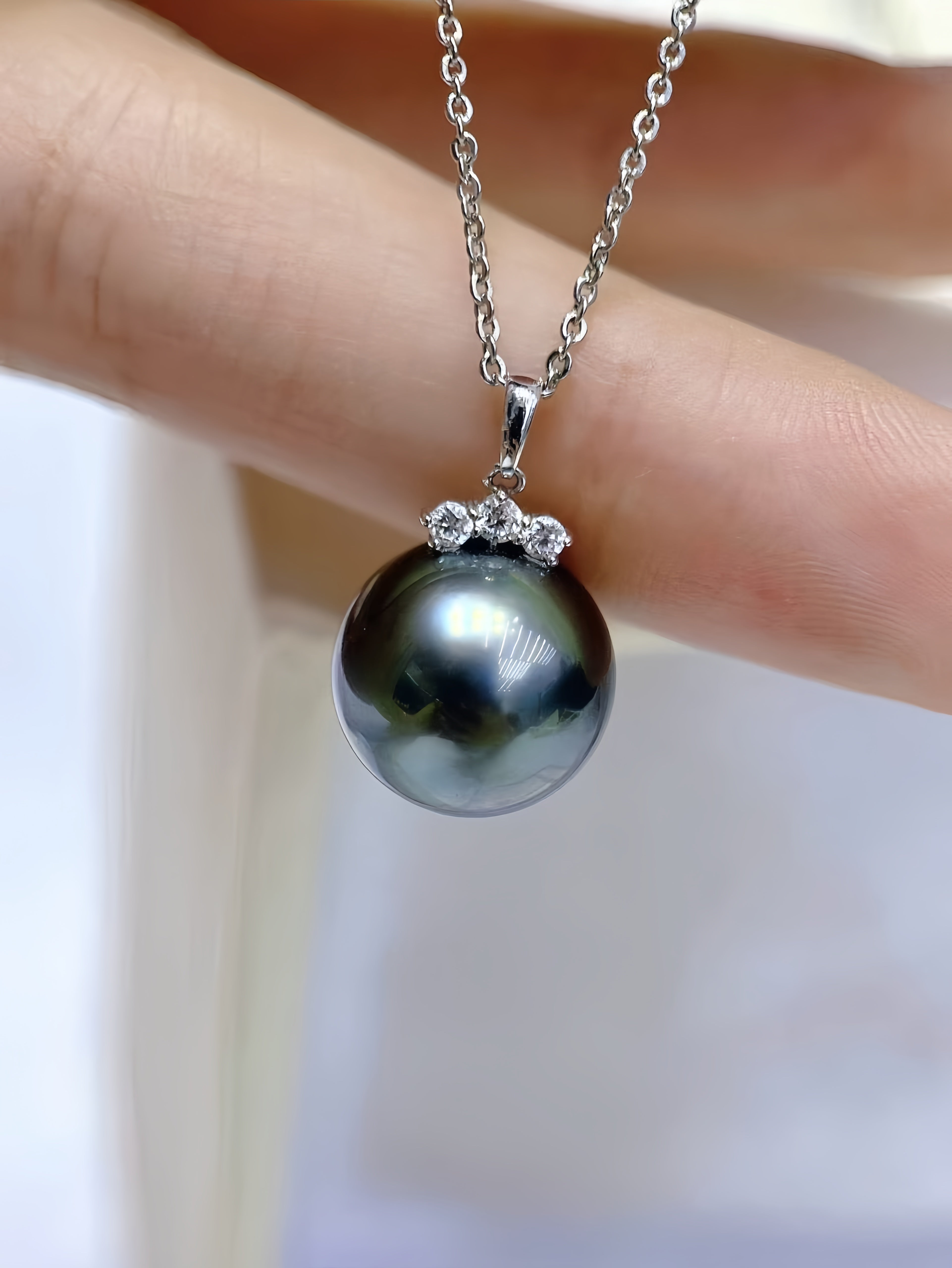 A close-up of a hand holding a silver pendant necklace featuring a large, dark, iridescent pearl with small clear stones at the top, on a silver chain.