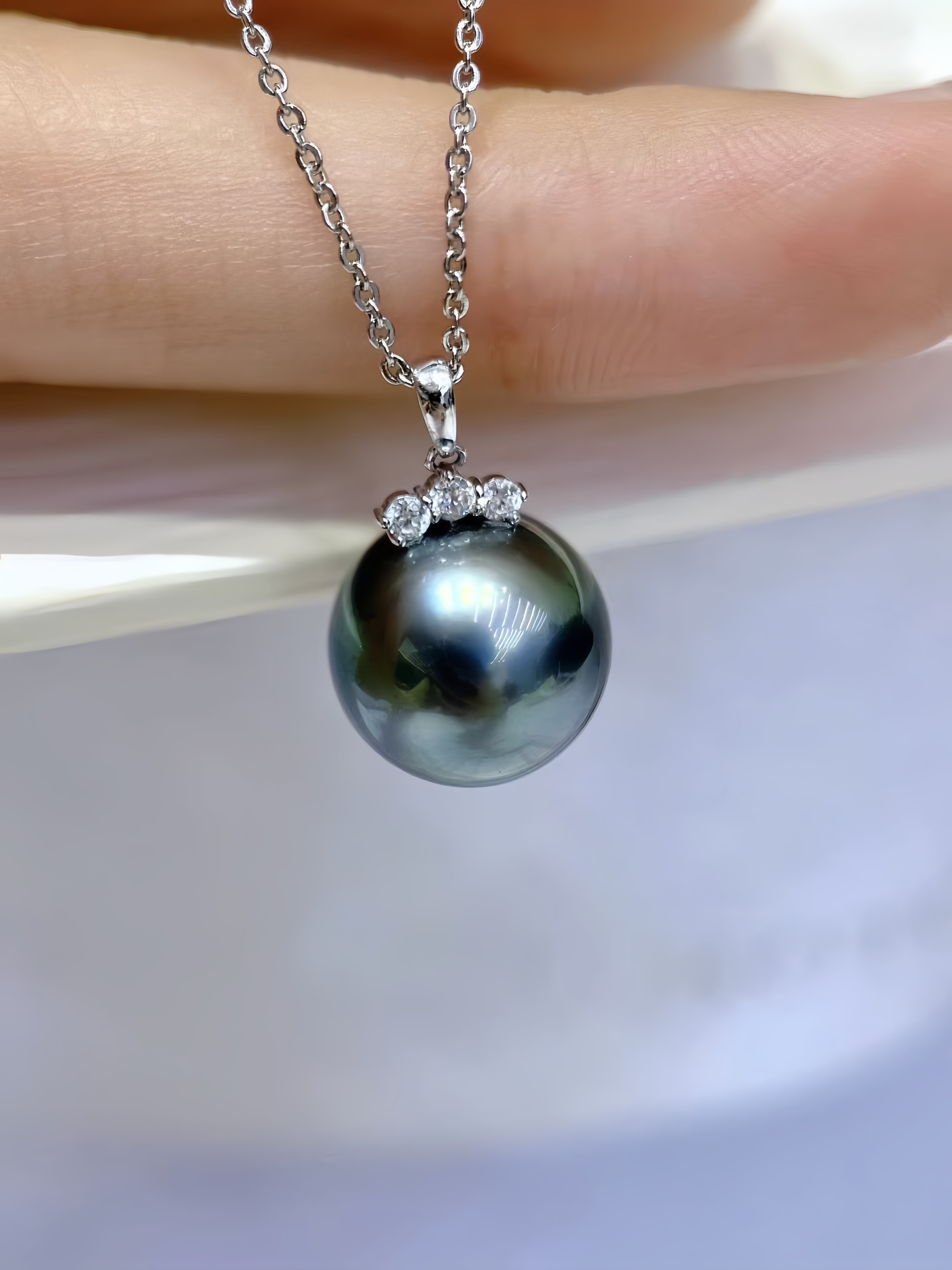 A close-up of a silver pendant necklace featuring a large, dark, iridescent freshwater pearl accented by three small clear stones, held against a soft-focus background.