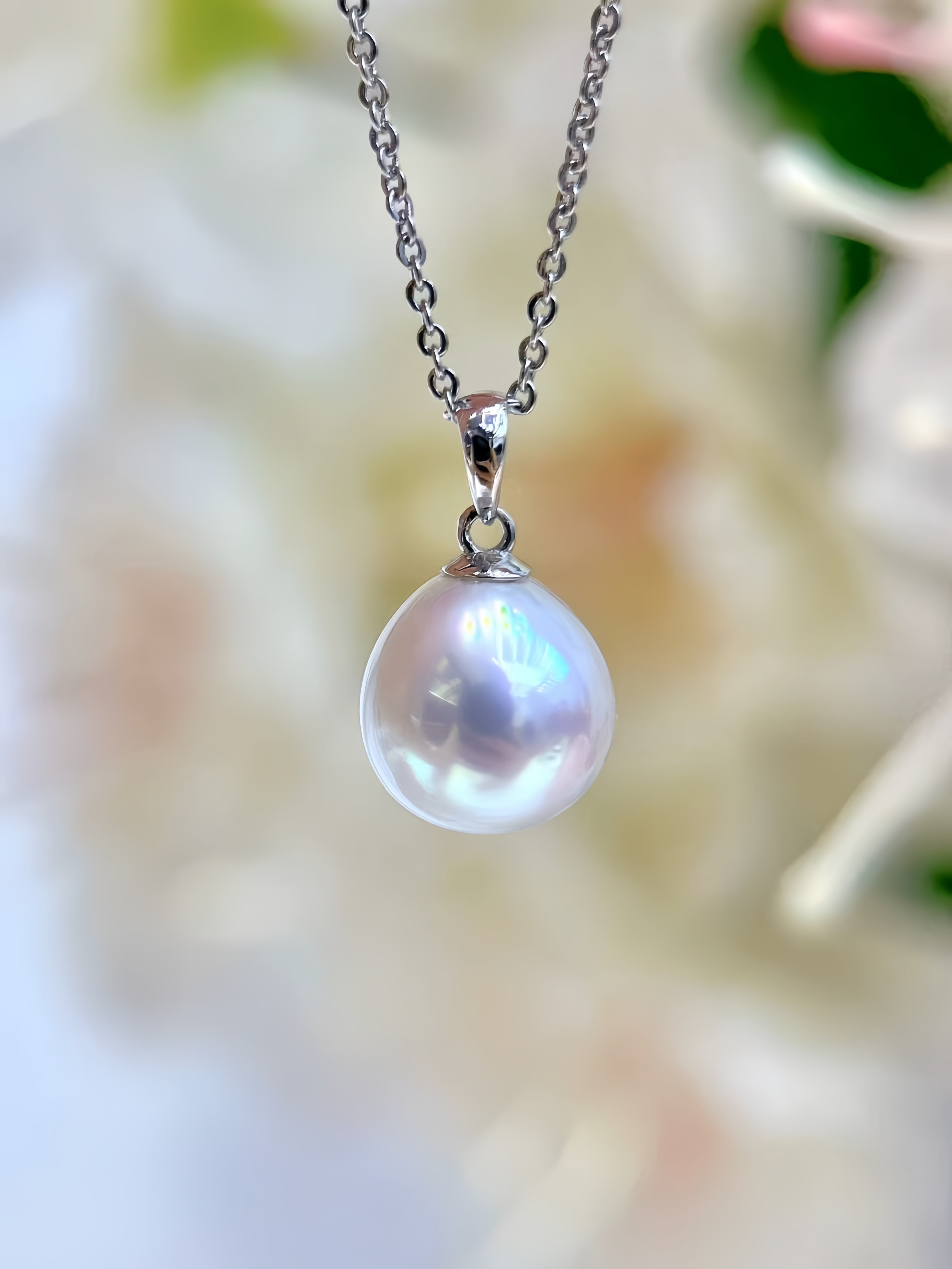 A close-up of a silver chain necklace with a teardrop-shaped white pearl pendant, set against a soft, blurred background of white flowers and green leaves.