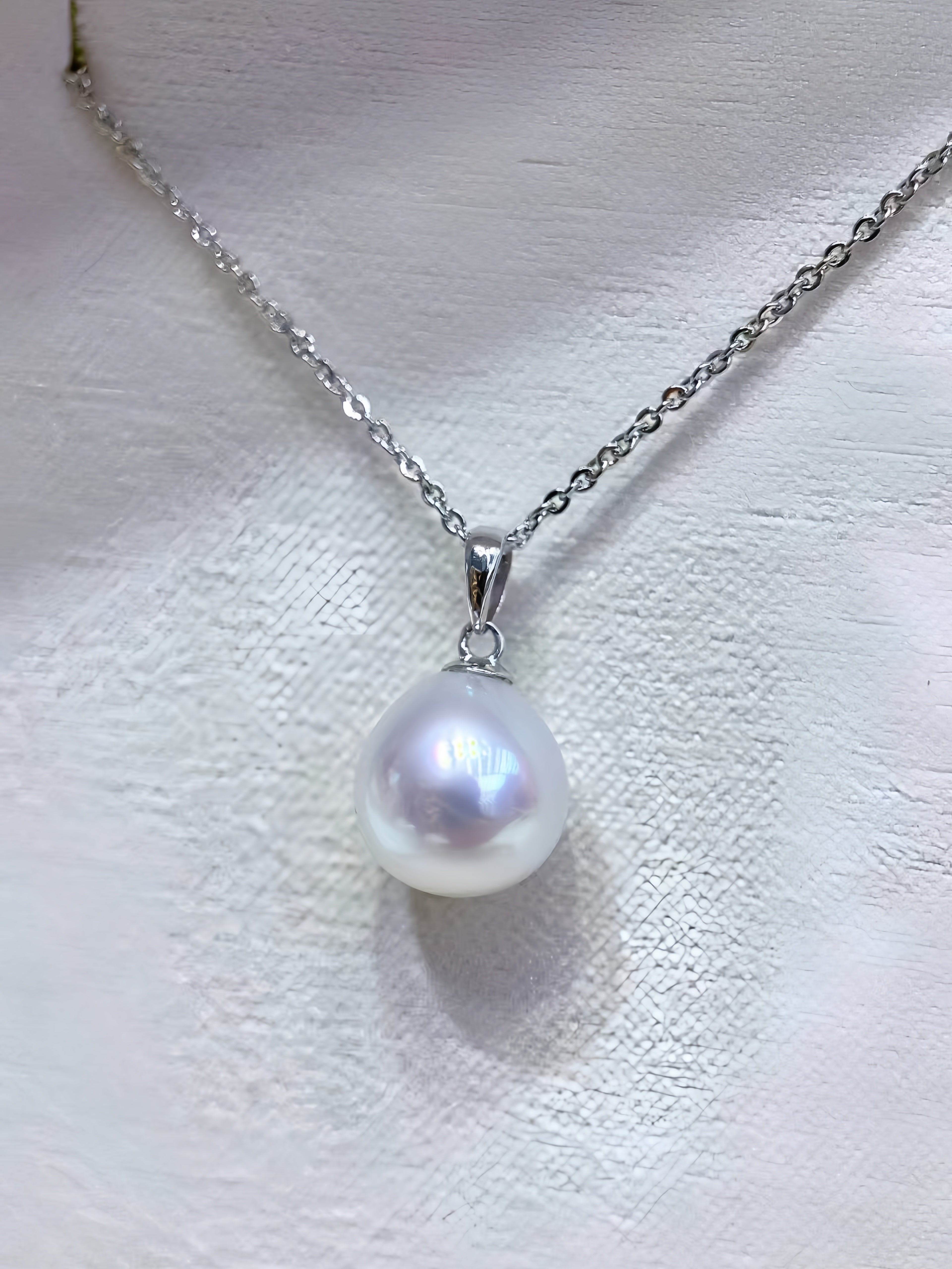 A silver chain necklace with a single teardrop-shaped white pearl pendant, displayed on a textured white surface.