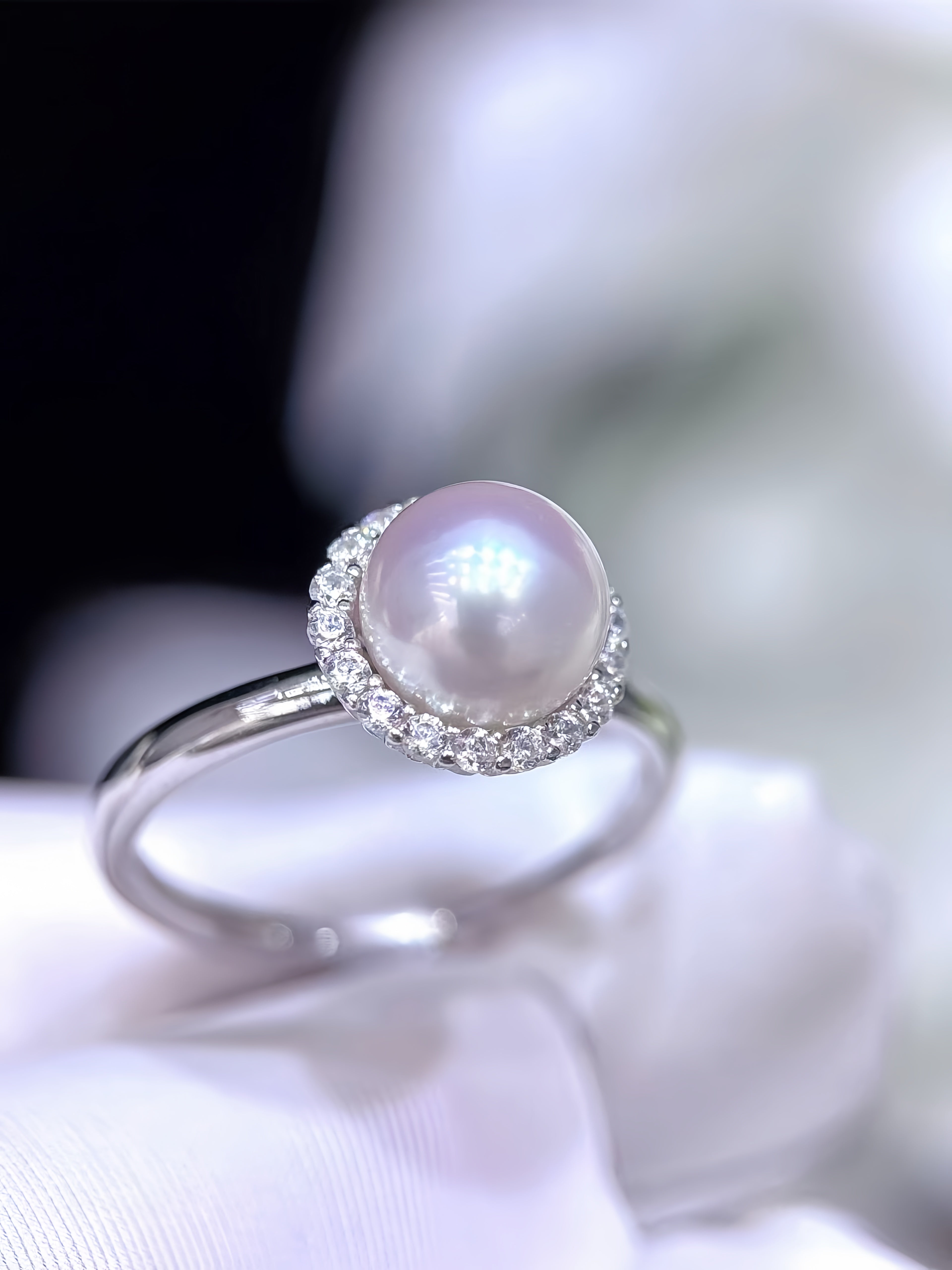 A silver ring featuring a large, lustrous white pearl surrounded by a halo of small clear stones, resting on a soft white fabric surface.