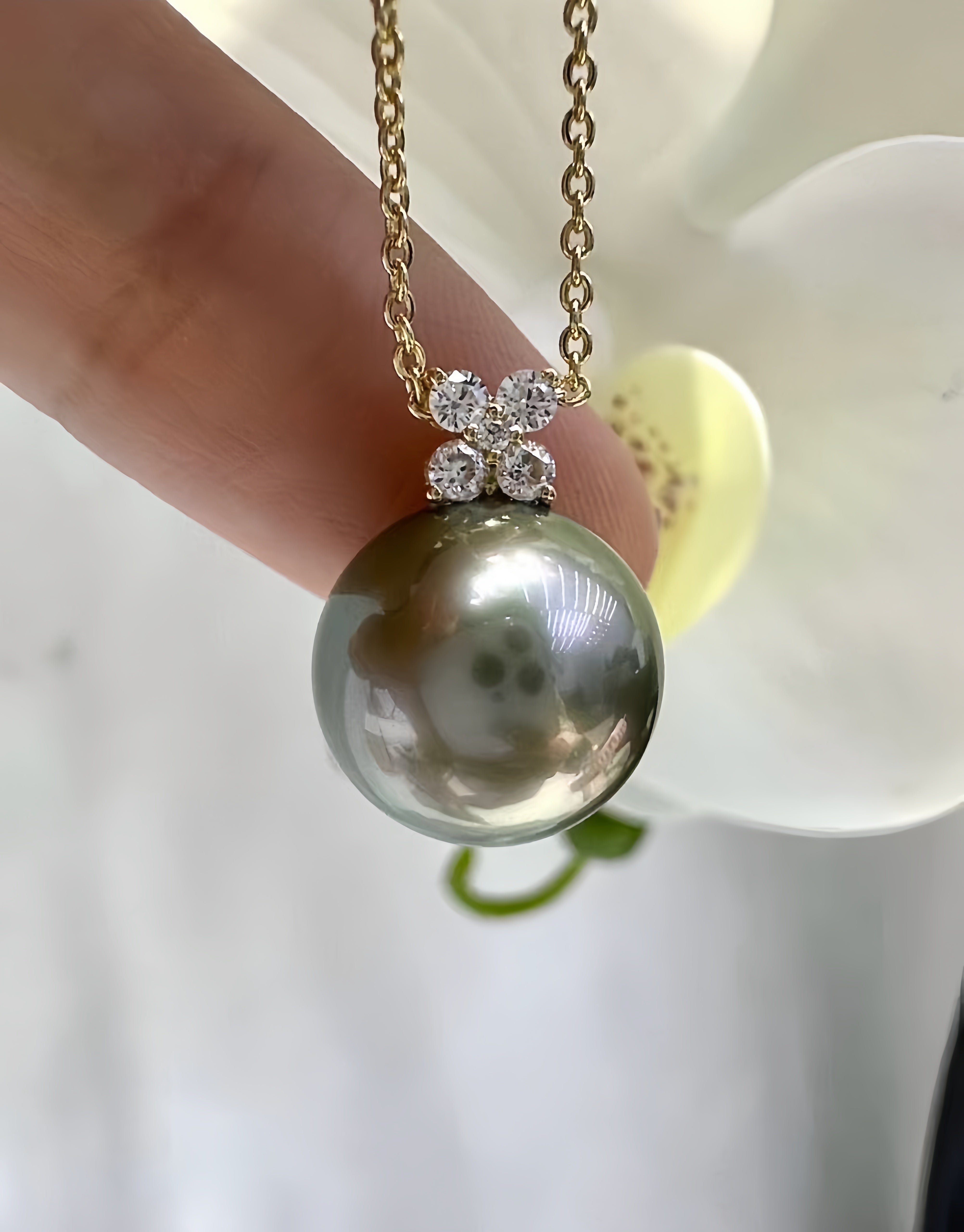 A close-up of a gold chain necklace with a large round gray pearl pendant, held by a finger in front of a white flower.