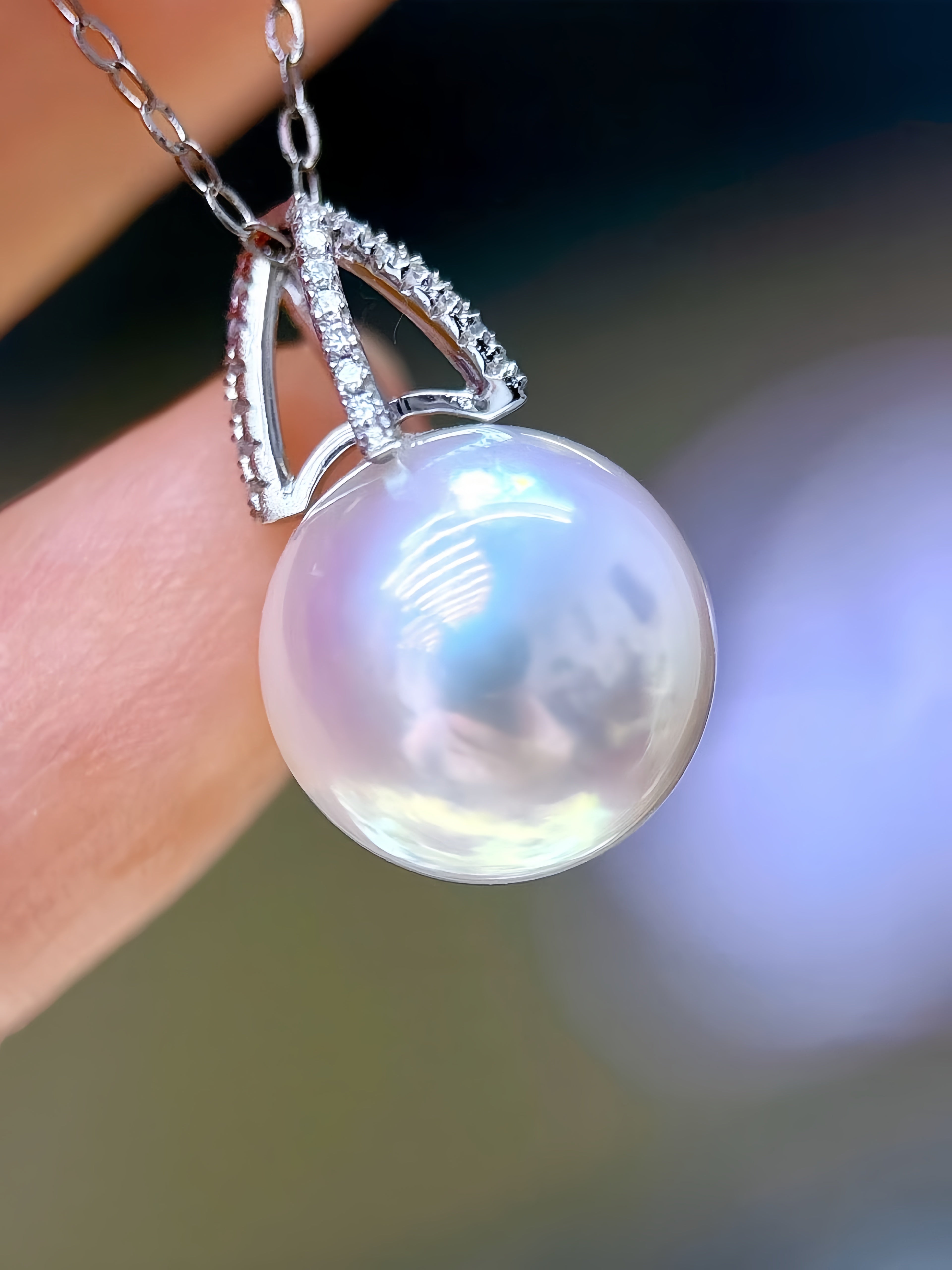 A close-up of a silver pendant necklace with a large, lustrous white pearl and small clear stones, held by a person's fingers against a blurred background.
