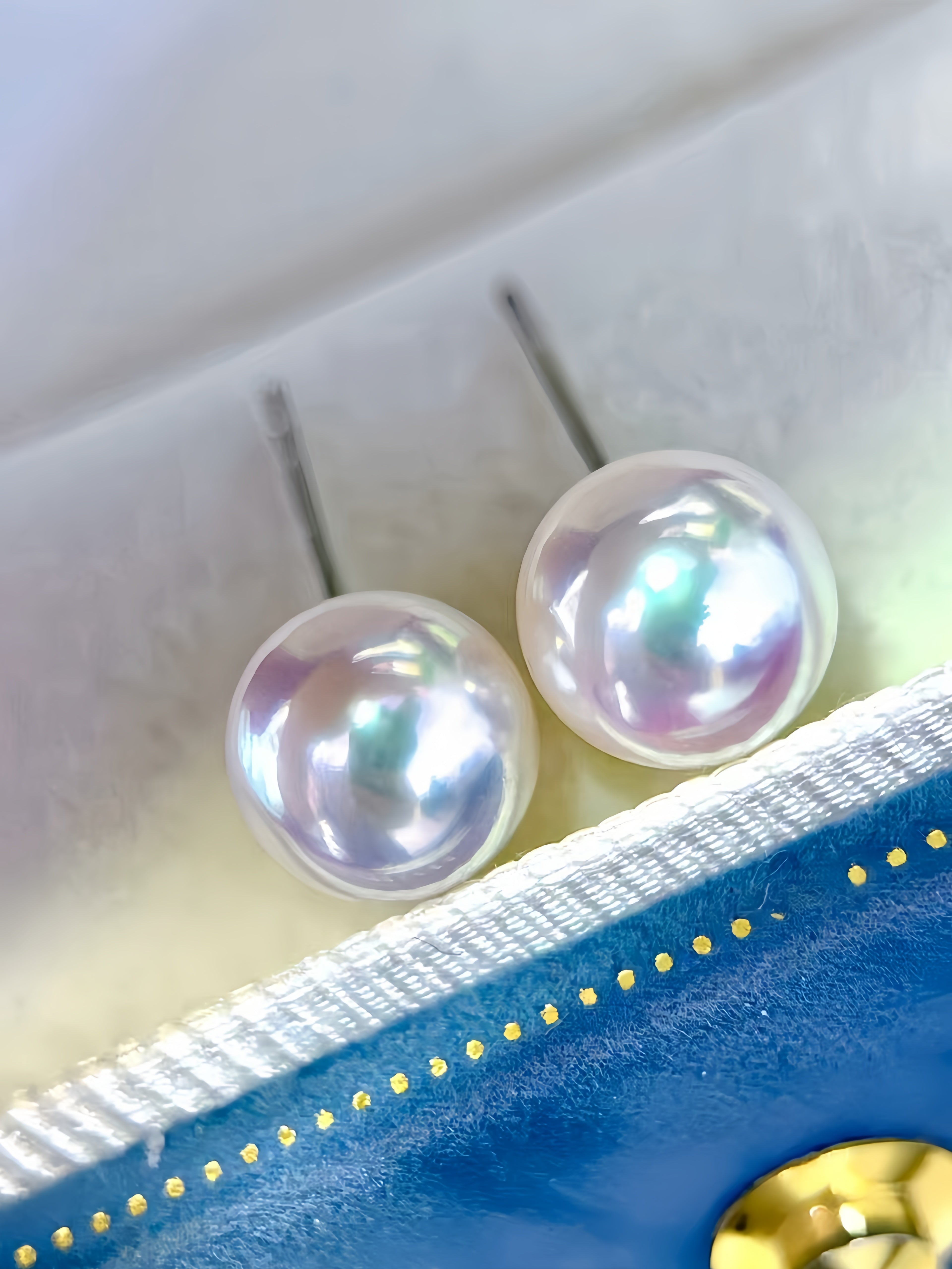 A pair of round lustrous pearl stud earrings with silver posts, resting on a white textured surface near a blue and gold decorative prop.