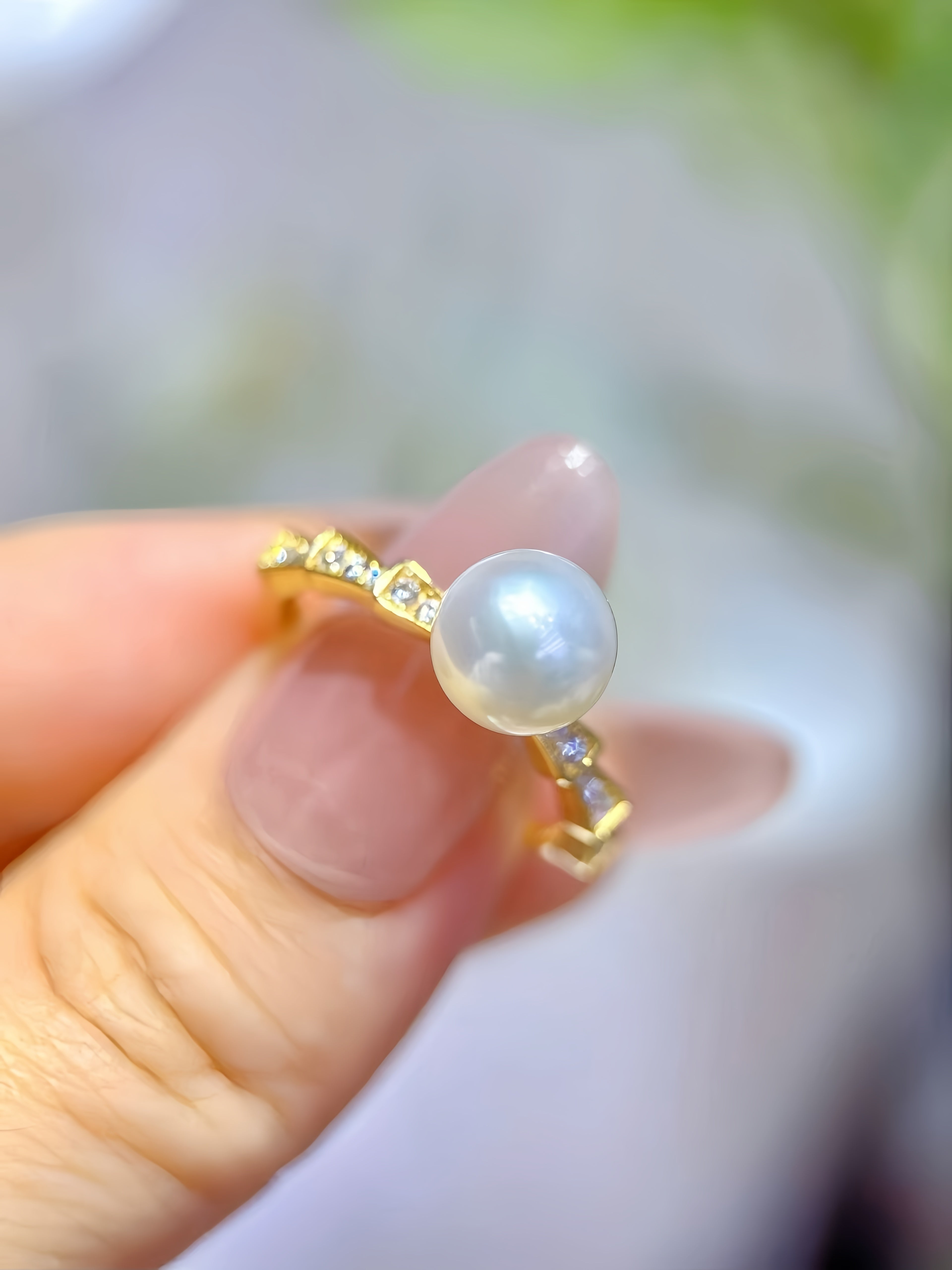 A gold ring with a large white pearl and small clear stones, held between fingers against a soft blurred background.