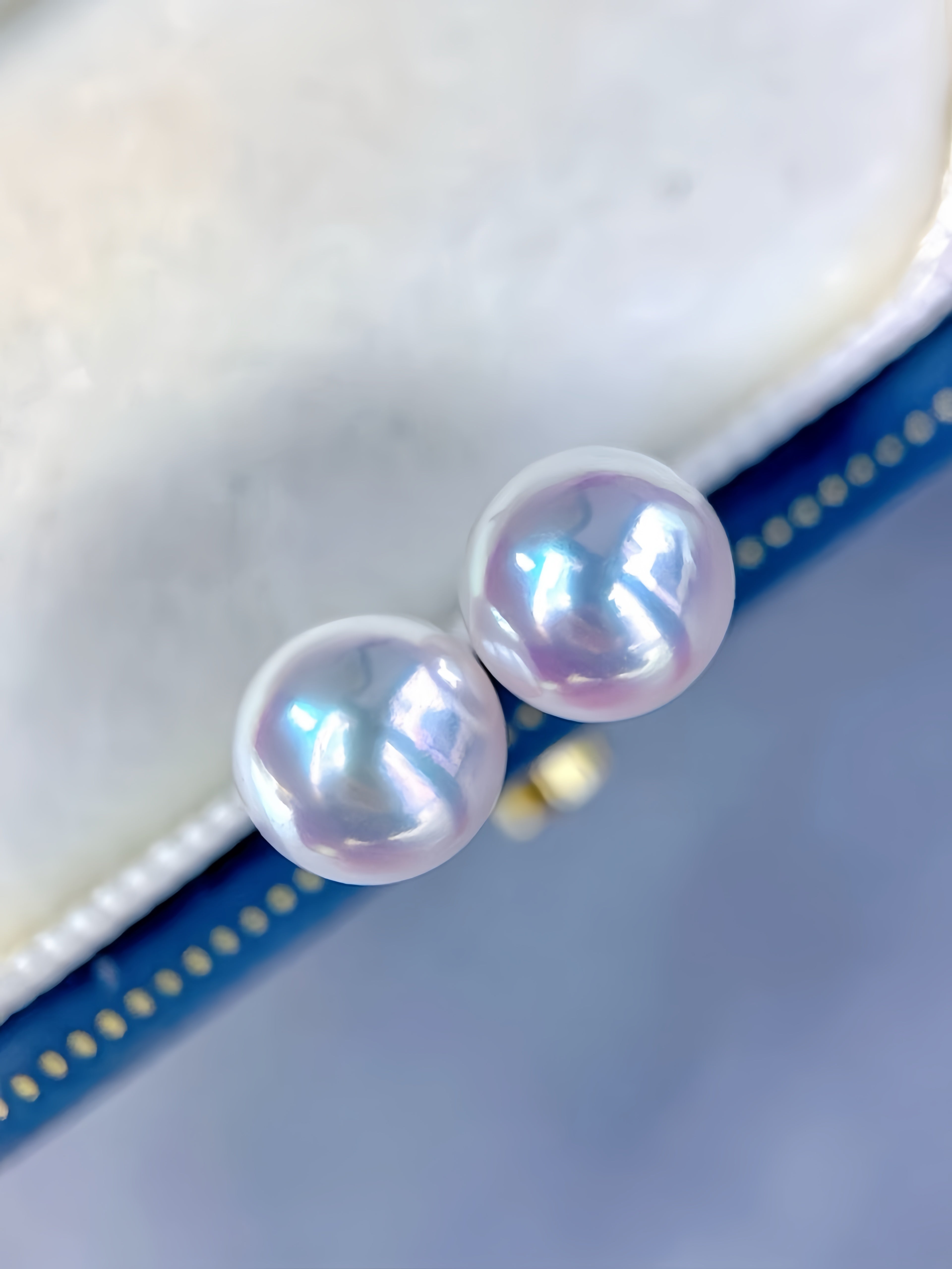 Two round lustrous pearl stud earrings with a soft iridescent lavender-pink sheen, resting on a cream jewelry box lid over a blue surface.