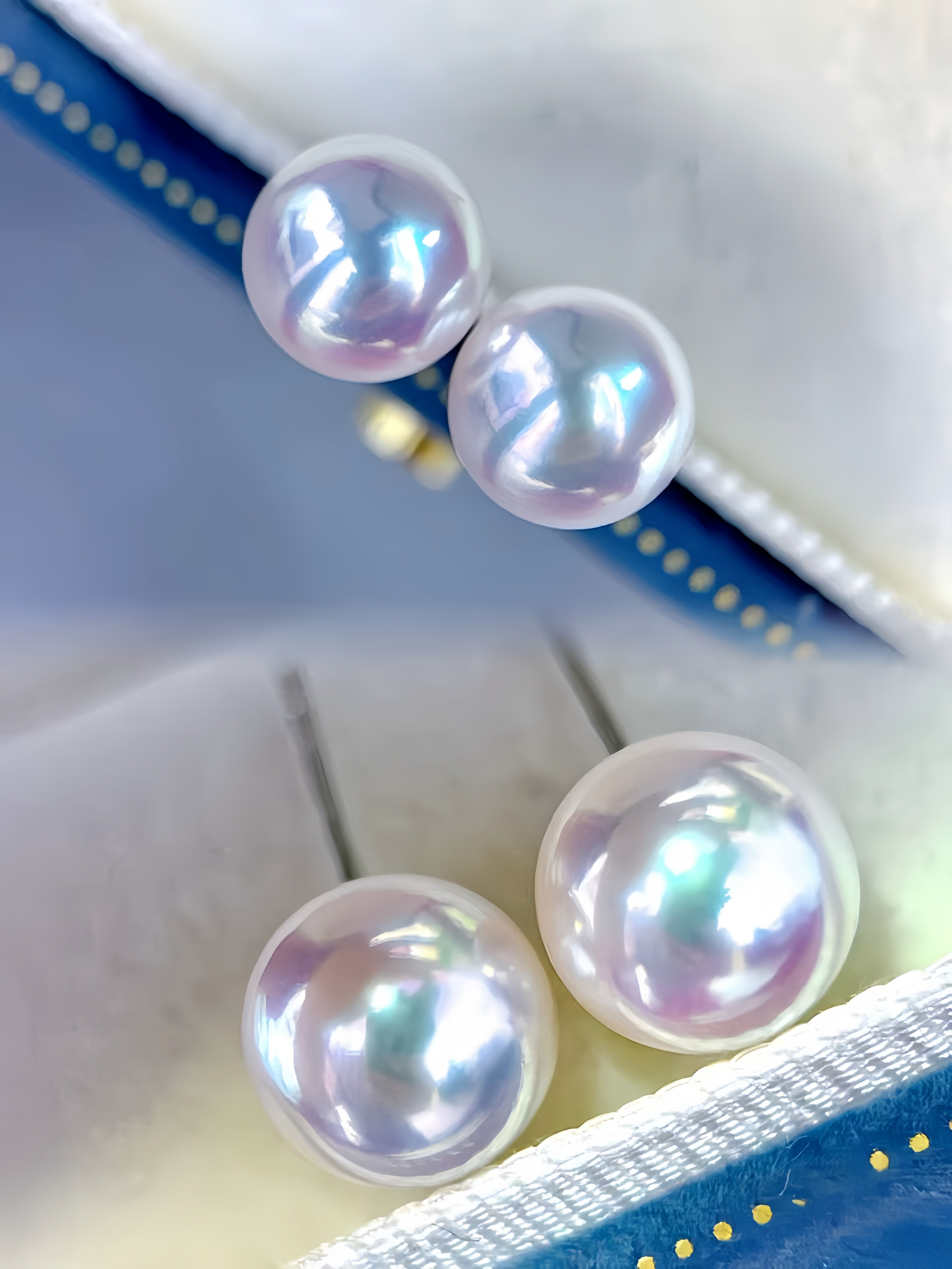 Three round iridescent pearl stud earrings displayed on a soft cream jewelry box with blue ribbon accents.