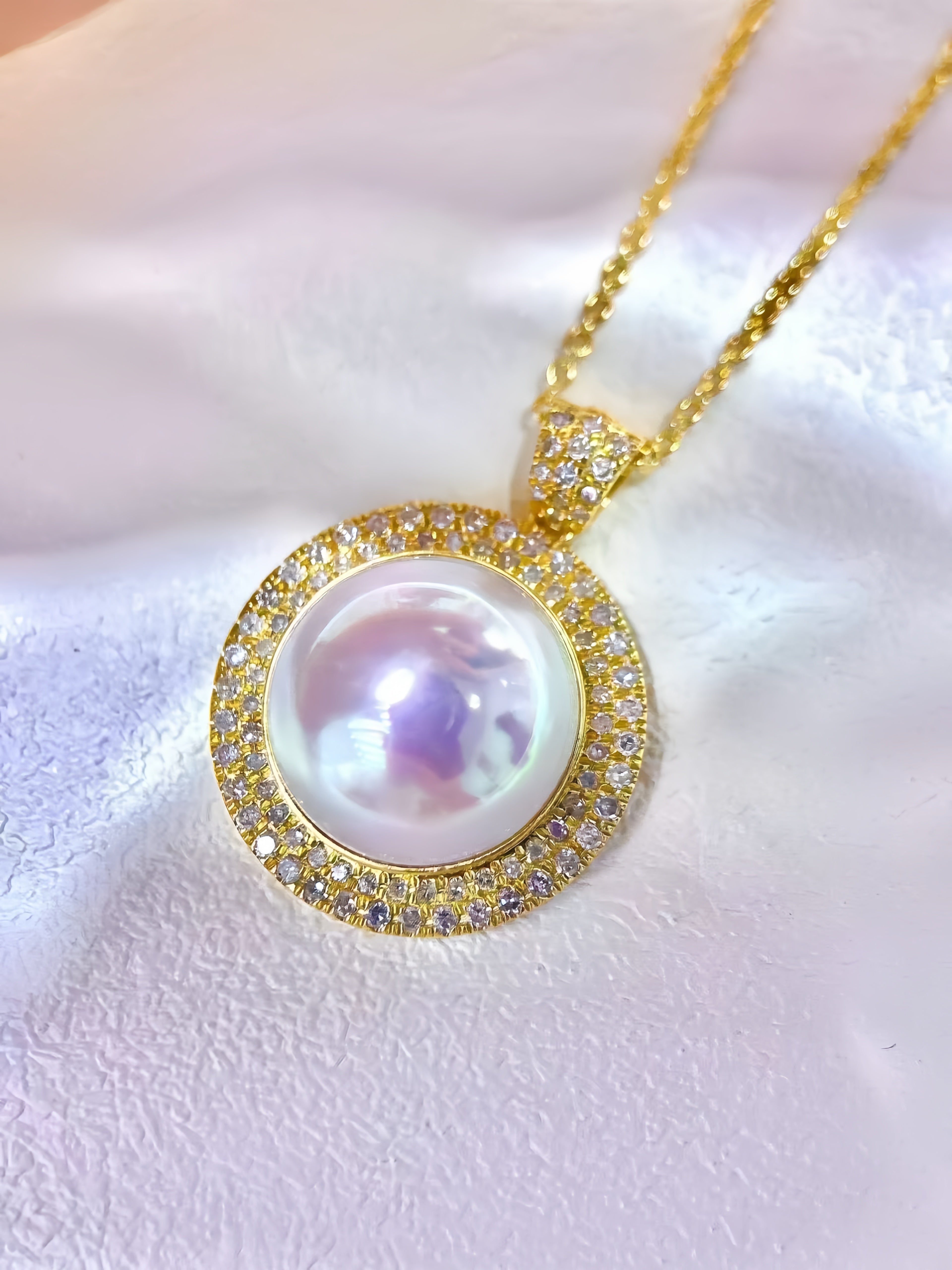 A gold pendant necklace featuring a large iridescent white pearl surrounded by a halo of small clear stones, displayed on a soft white textured fabric background.
