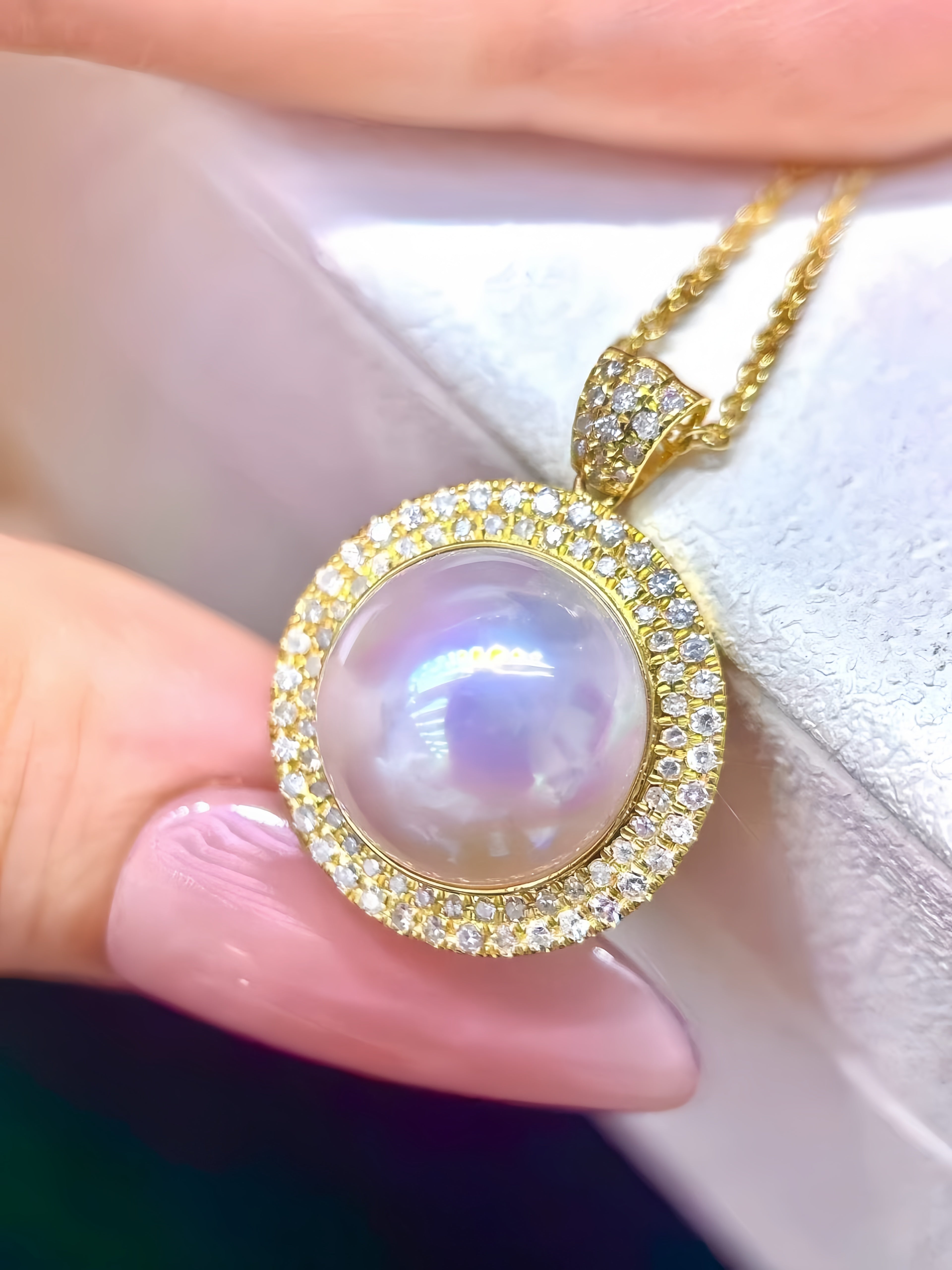 A gold pendant necklace with a large iridescent pearl surrounded by a halo of small clear stones, held by a hand against a soft white background.