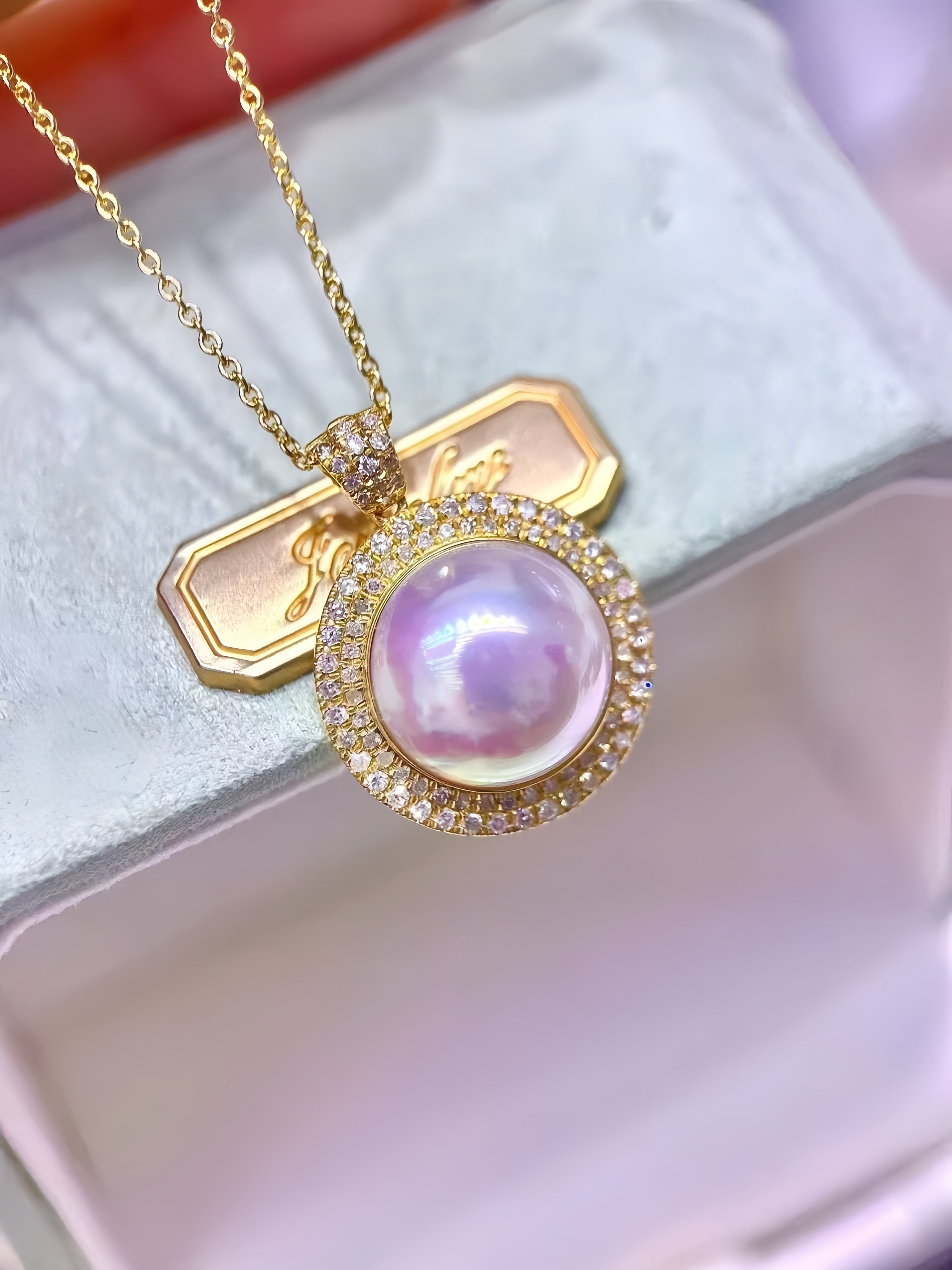 A gold pendant necklace featuring a large iridescent pink pearl surrounded by small clear stones, resting on a white jewelry box.