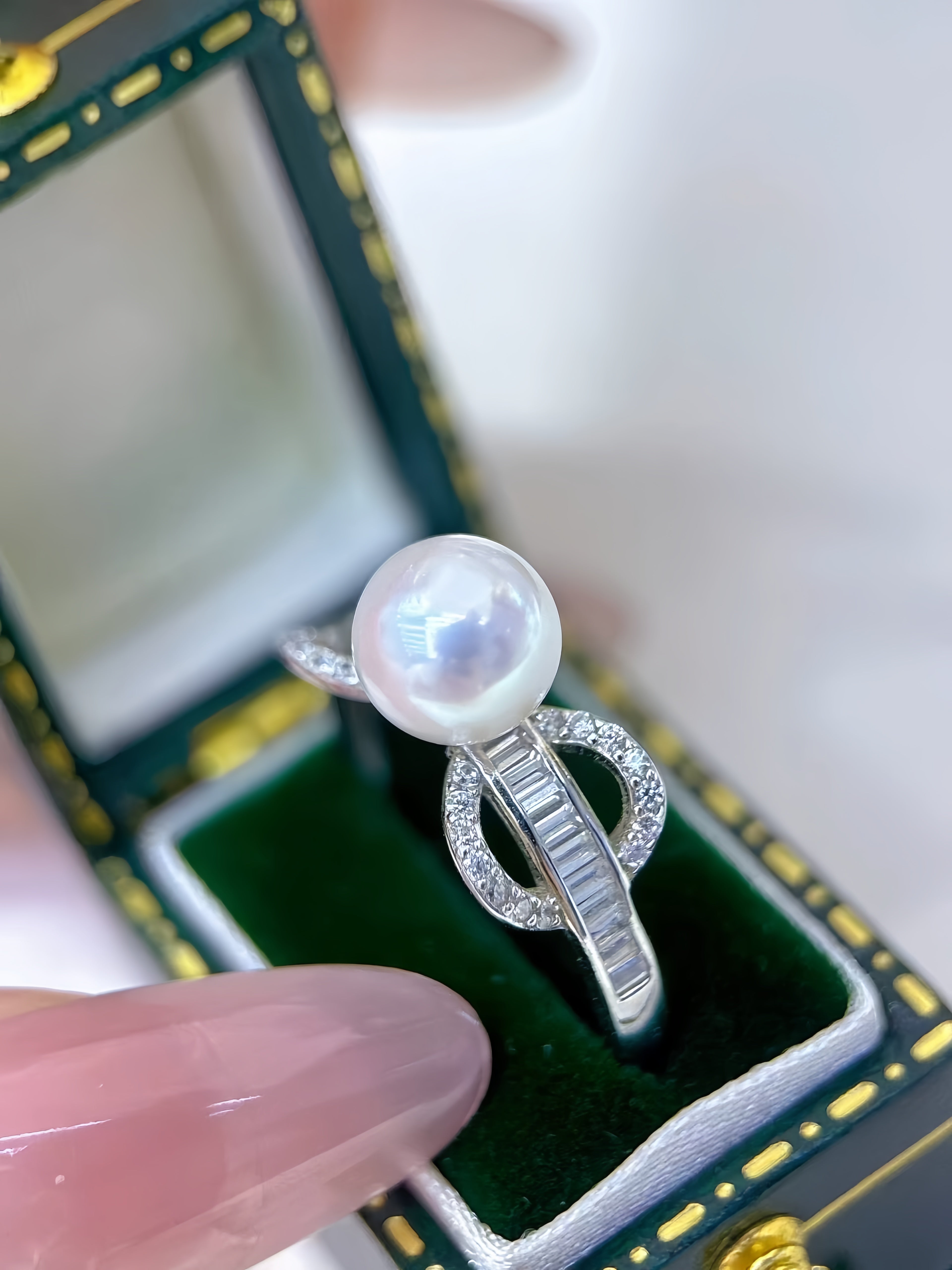 A silver ring with a large lustrous pearl and baguette-cut stones, displayed in an open dark green jewelry box with a finger holding it.