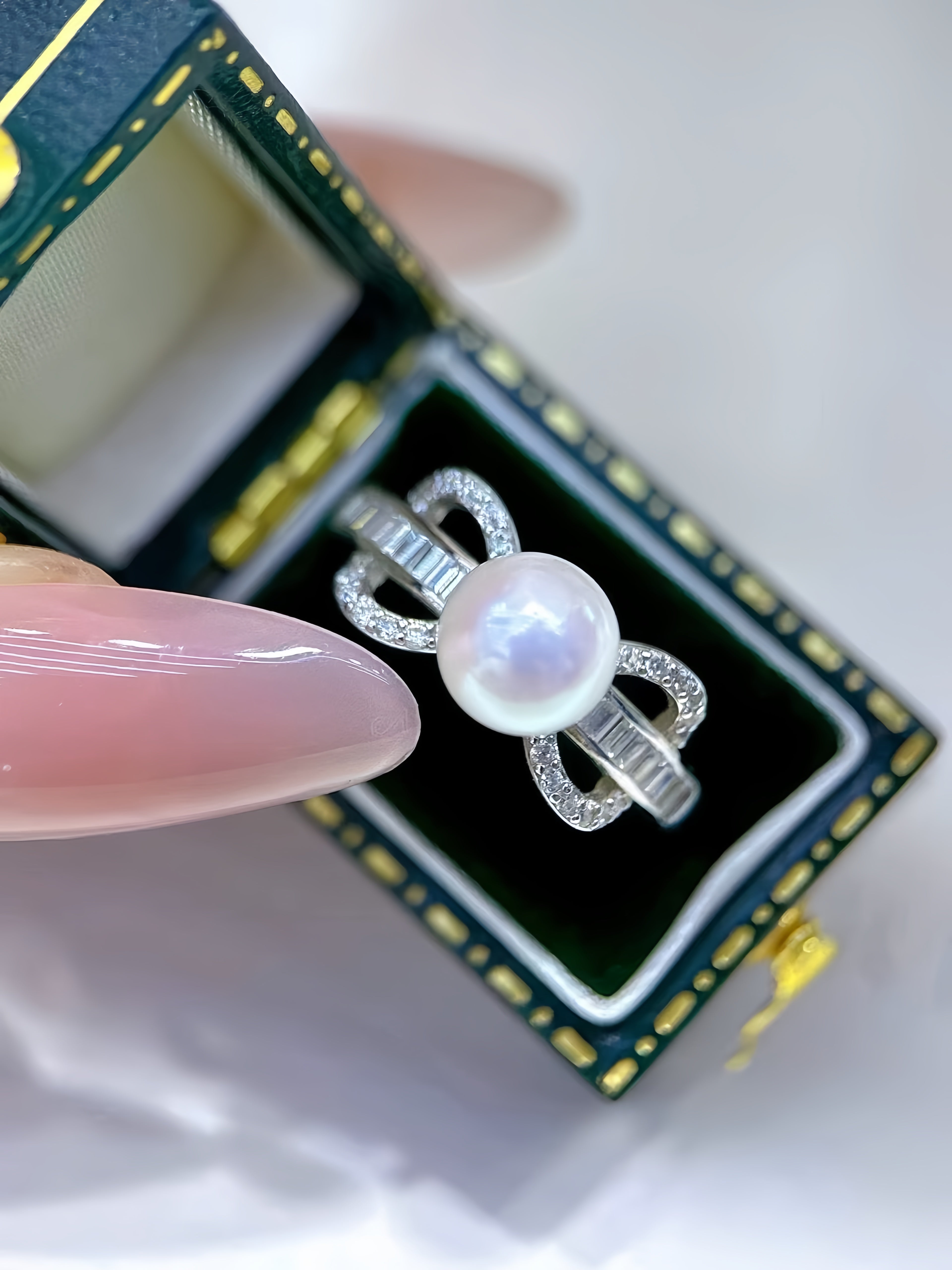 A silver ring with a large round white pearl and small clear stones, held by a manicured finger in front of an open dark green jewelry box.