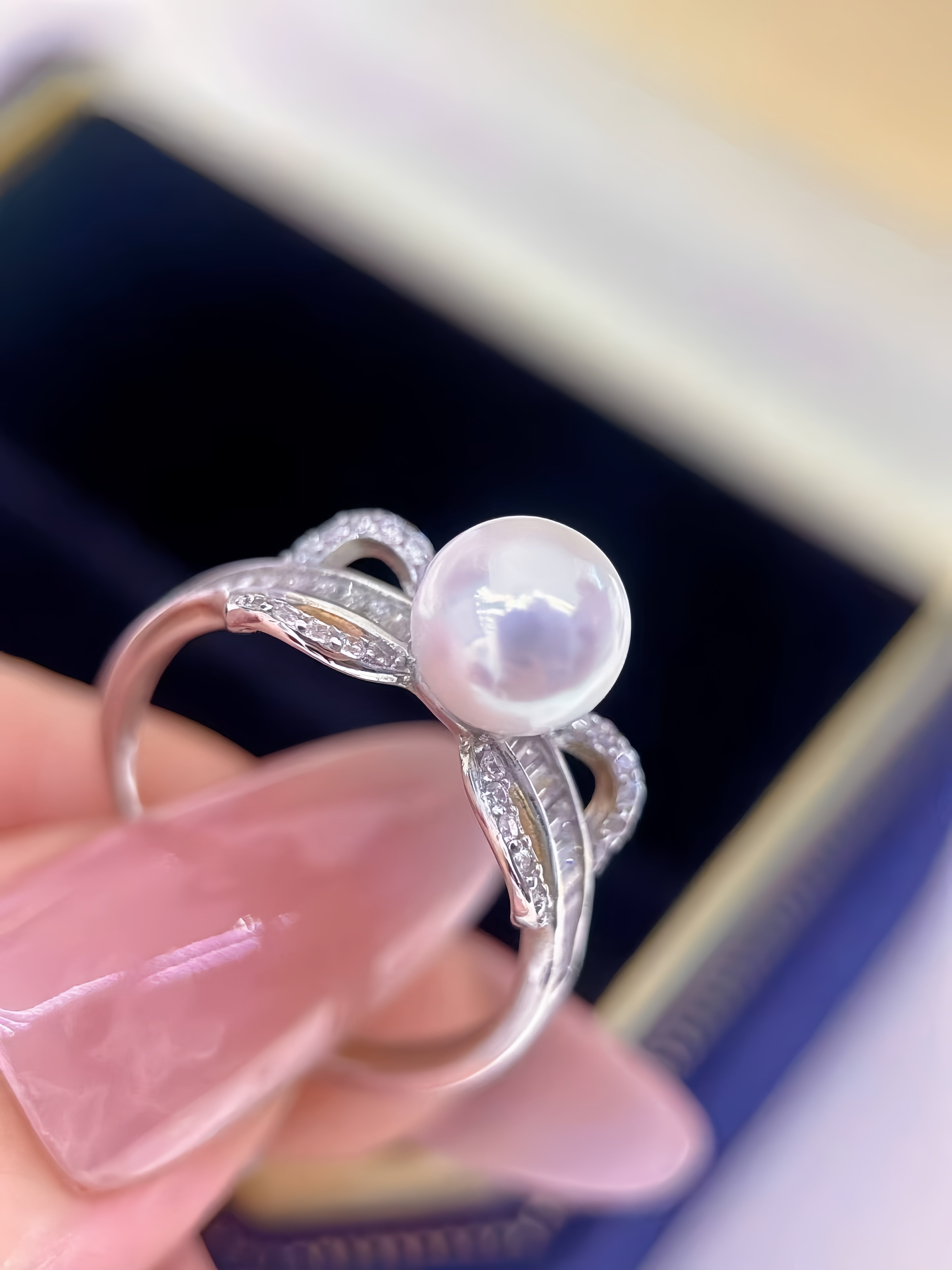 A silver ring with a round white pearl and small clear stones, held by a hand with pink manicured nails in front of a dark blue jewelry box.