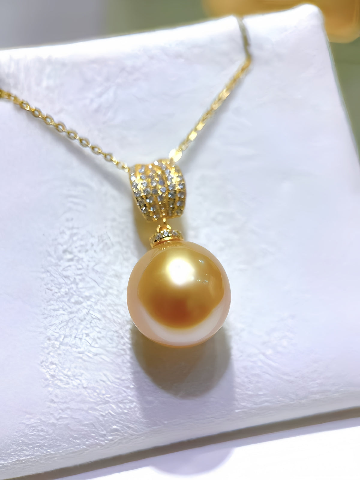 A gold pendant necklace featuring a large, lustrous golden pearl suspended from a gold chain with a pavé-set crystal accent, displayed on a textured white jewelry stand.