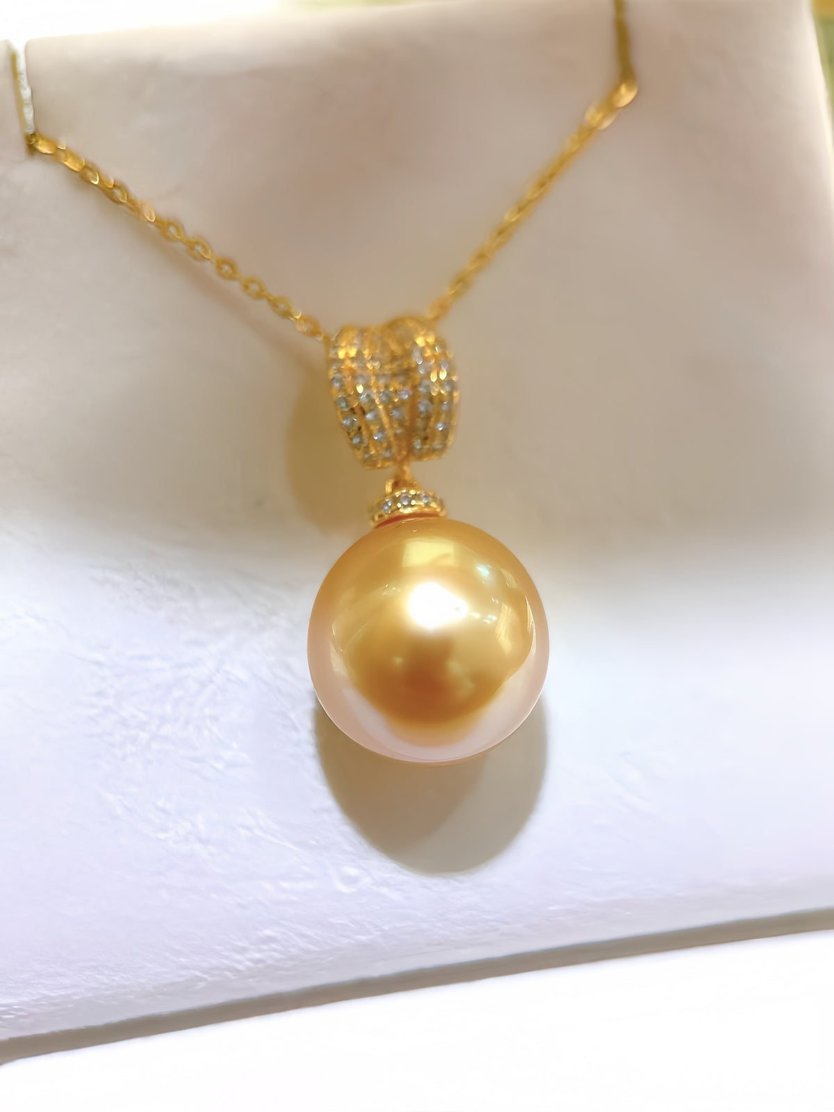 A gold pendant necklace featuring a large, round, iridescent gold pearl suspended from a gold chain with a pavé-set accent, displayed on a white jewelry stand.