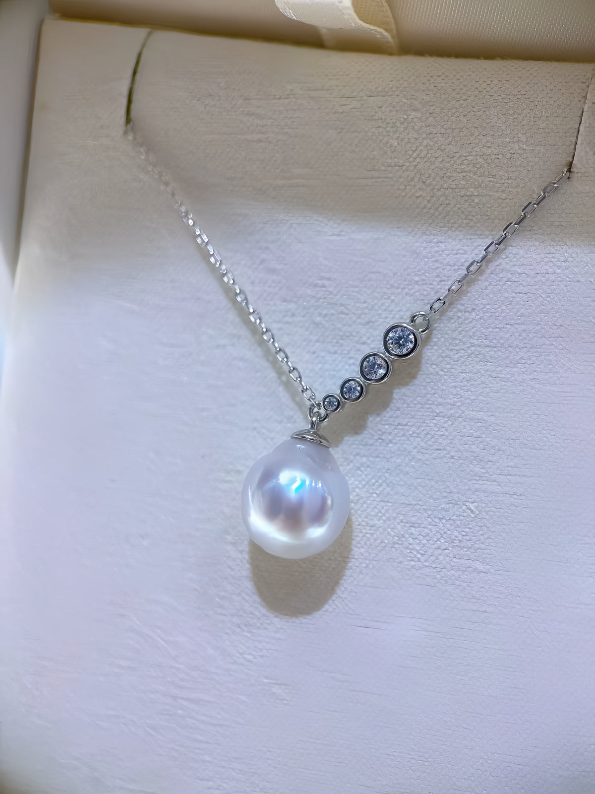 A silver necklace with a large white baroque pearl pendant and three small clear stones along the chain, displayed on a cream-colored jewelry box.
