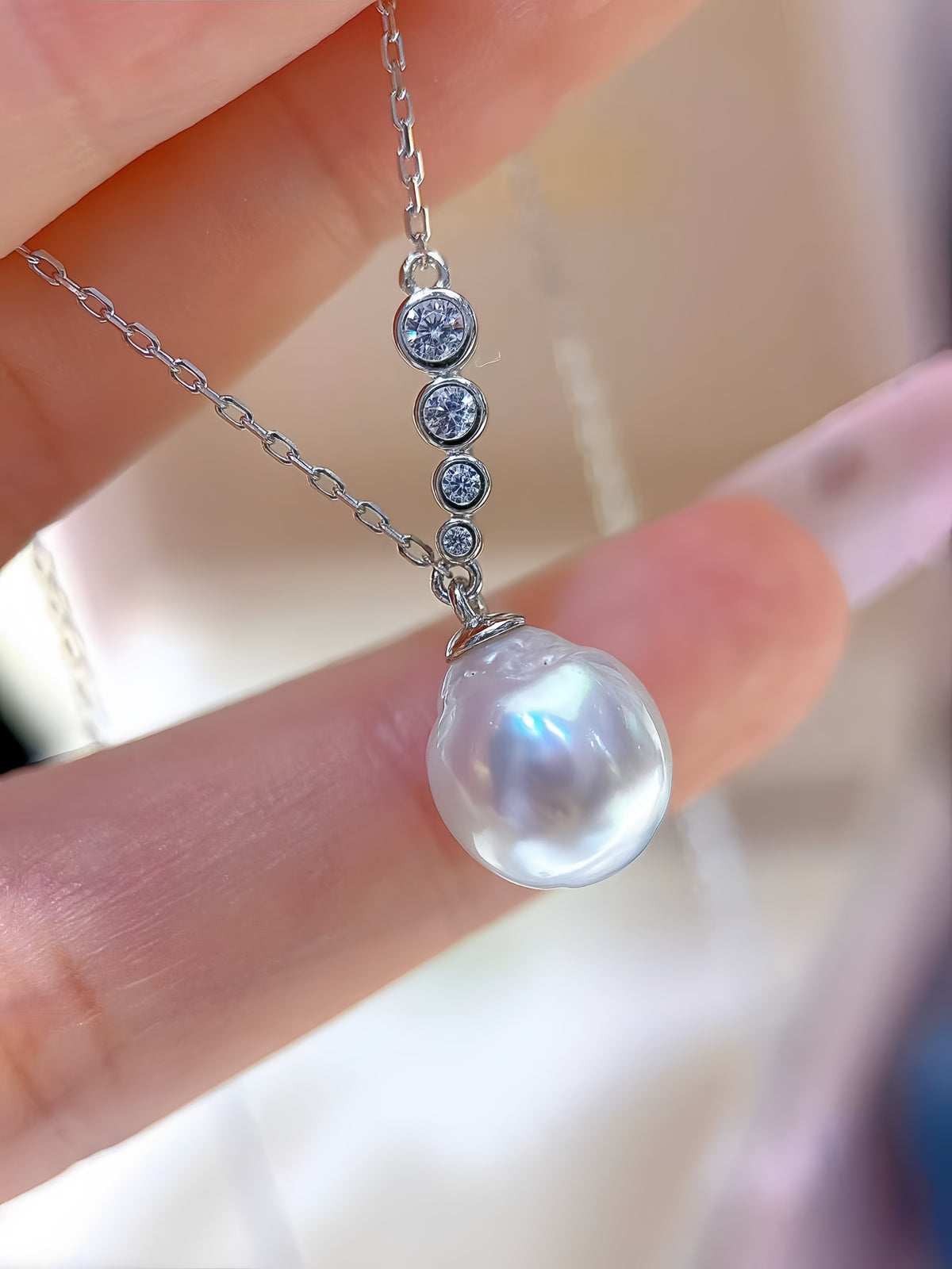 A close-up of a hand holding a silver necklace with a large, lustrous white pearl pendant and three small round clear stones above it.