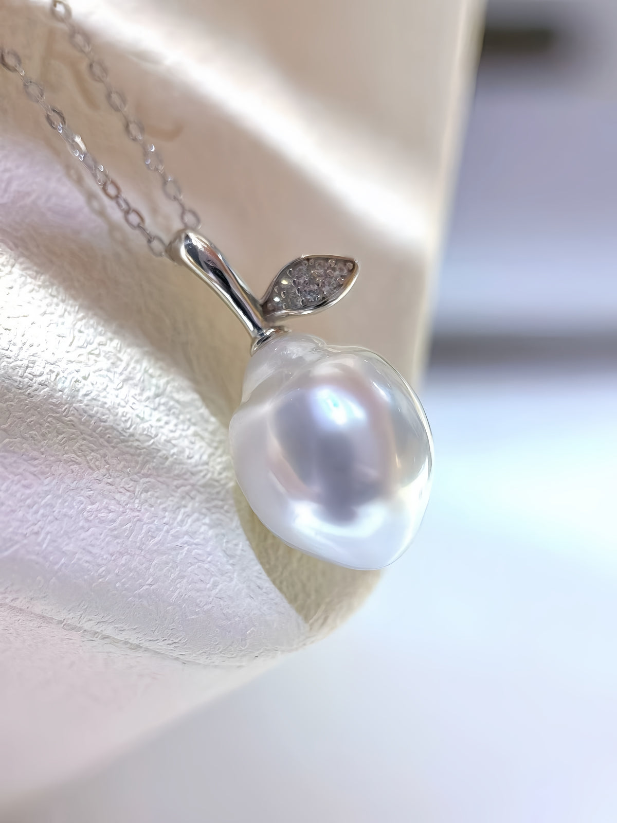 A silver pendant necklace shaped like an apple, featuring a lustrous white baroque pearl and a small leaf detail with tiny clear stones, displayed on a cream-colored jewelry stand.