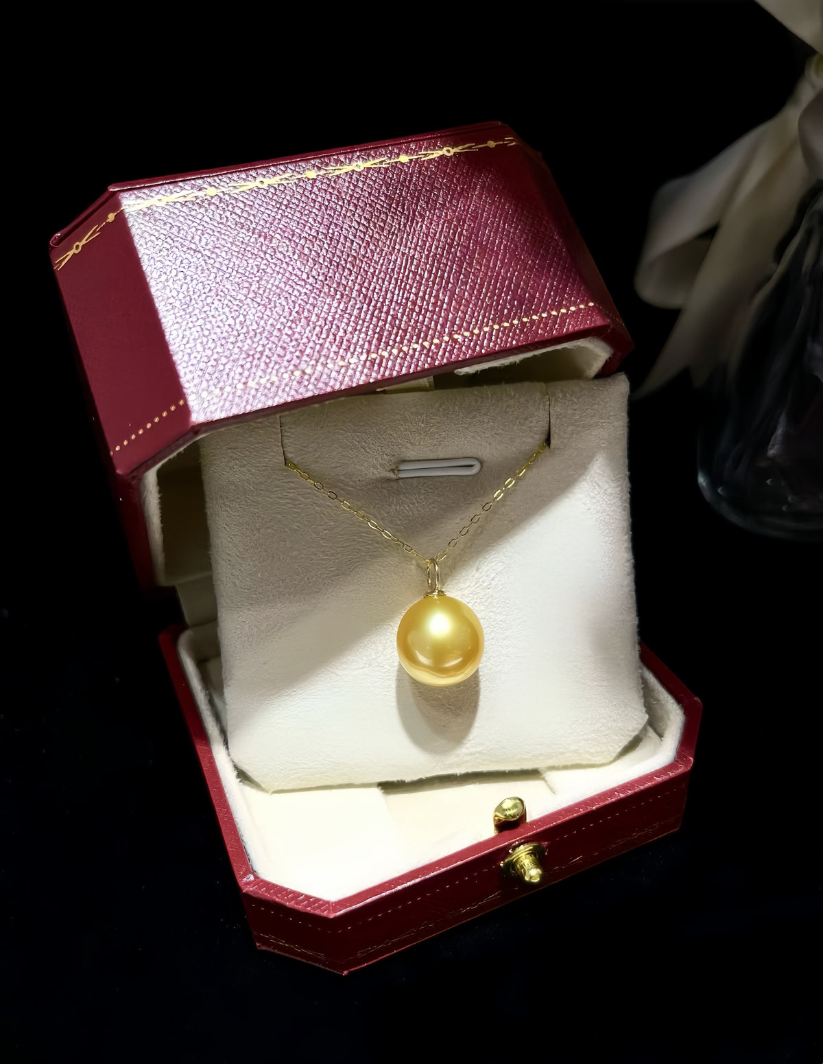 A gold pendant necklace with a round pearl-like drop is displayed inside an open red jewelry box with a cream insert, set against a dark background.