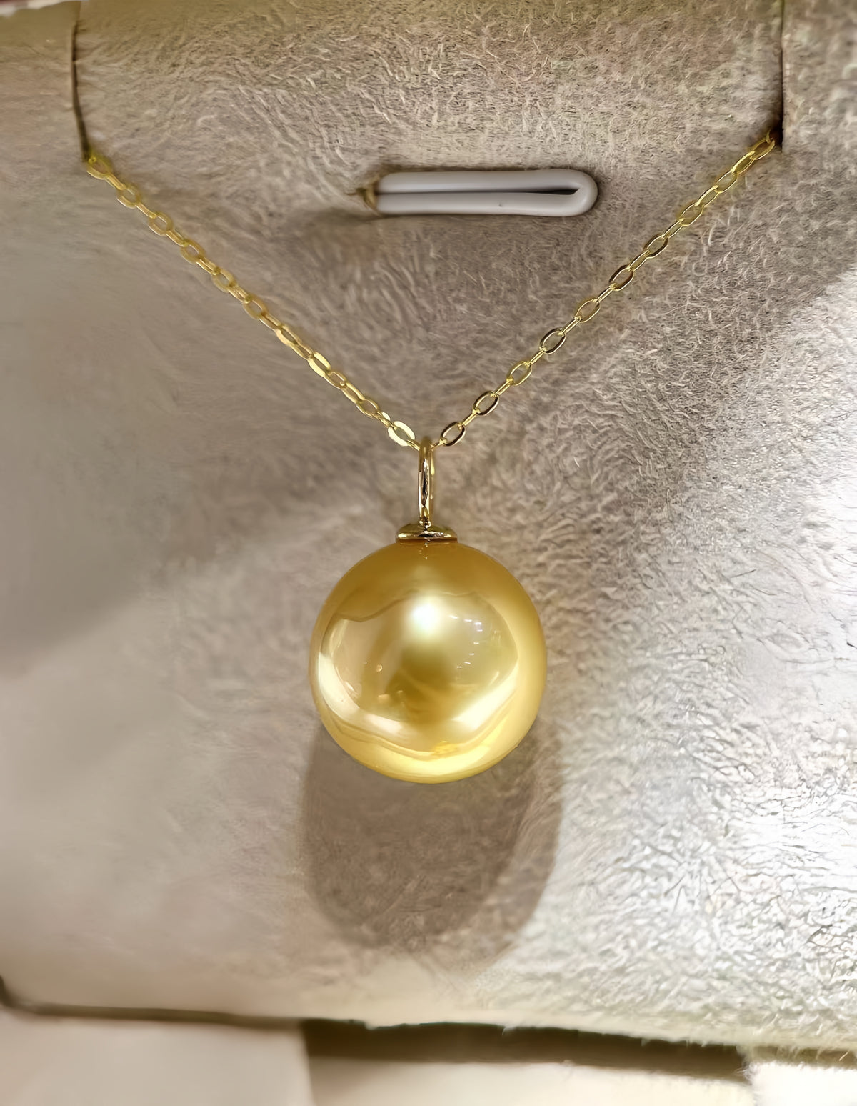 A gold chain necklace with a large, round, lustrous yellow pearl pendant, displayed on a textured cream-colored jewelry stand.