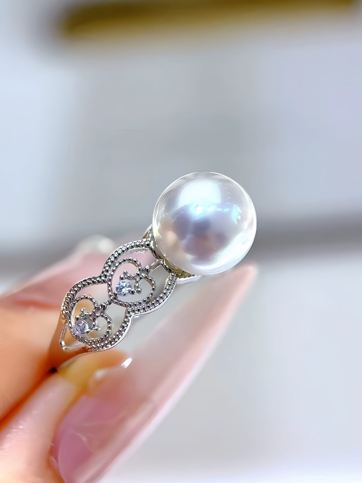 A silver ring with a large lustrous white pearl and small clear stones, shown being held on a person's fingers against a soft blurred light background.
