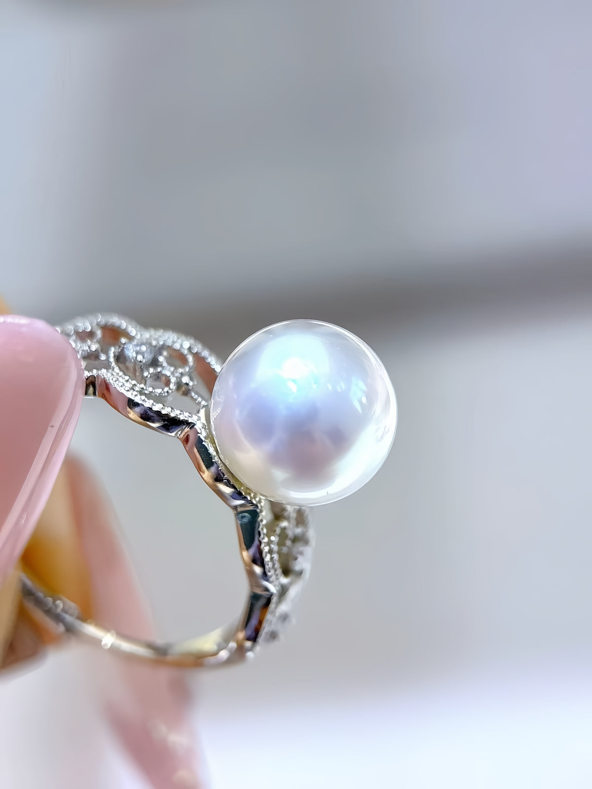A silver ring with an oval white pearl and ornate filigree detailing, held by a hand against a soft, out-of-focus light gray background.