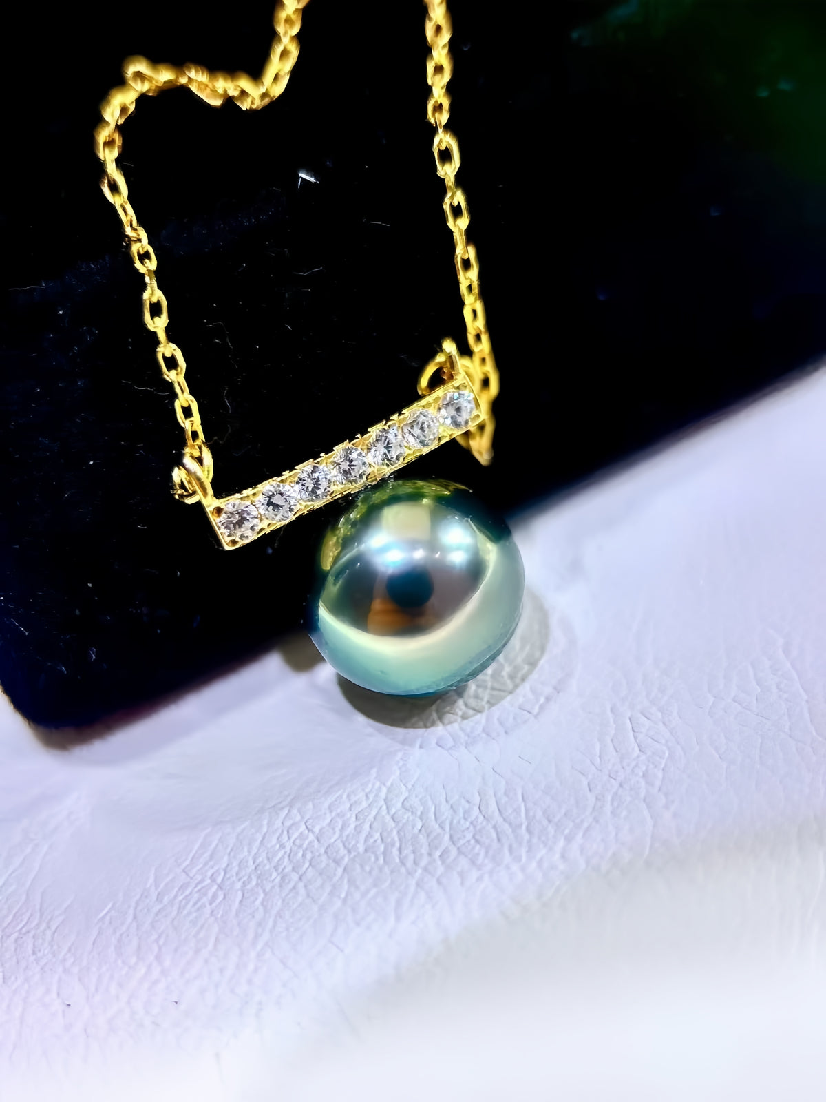 A gold necklace with a bar-set crystal pendant and a large round iridescent pearl pendant, displayed on a black jewelry stand against a white textured surface.