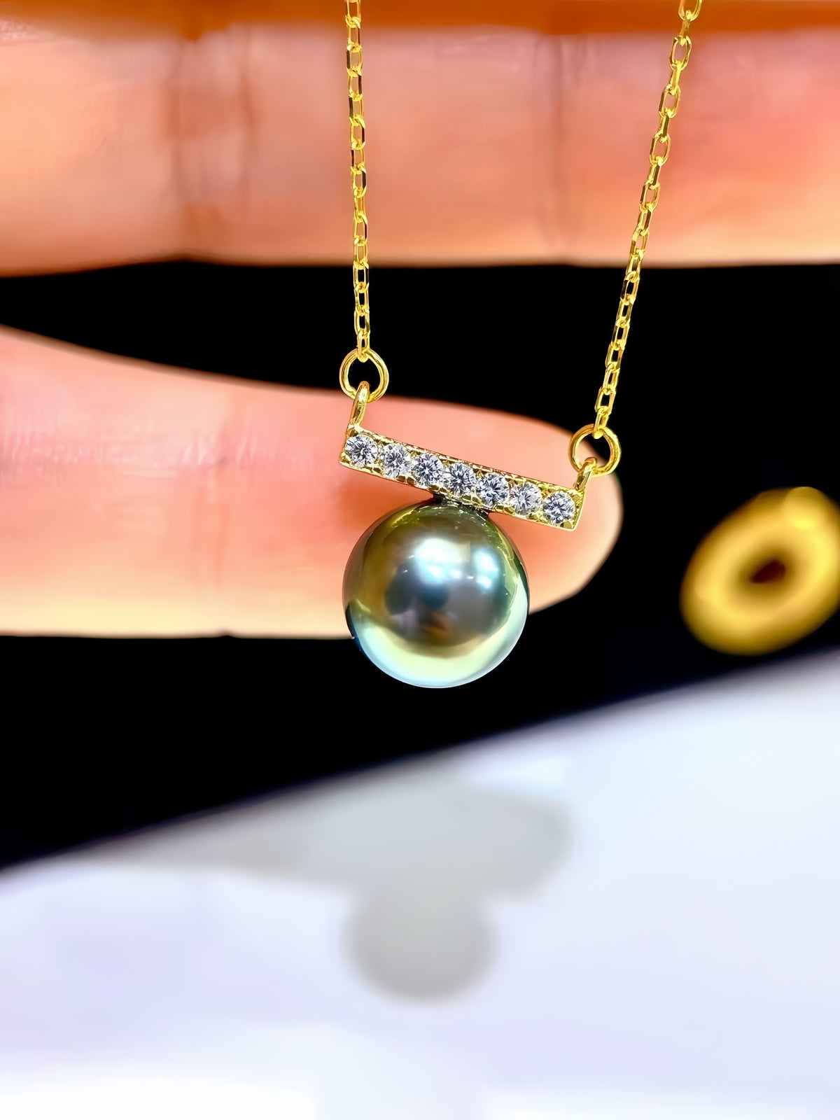 A gold necklace with a bar pendant set with small clear stones and a single round dark green pearl, held between fingers against a black and white background.