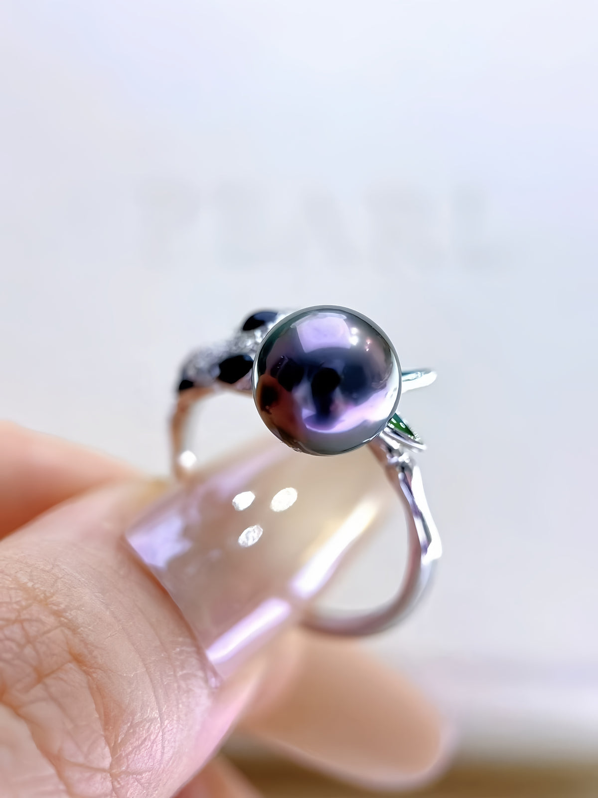 A silver ring with a large round black pearl and small green leaf accents, held on a person's finger against a soft white background.